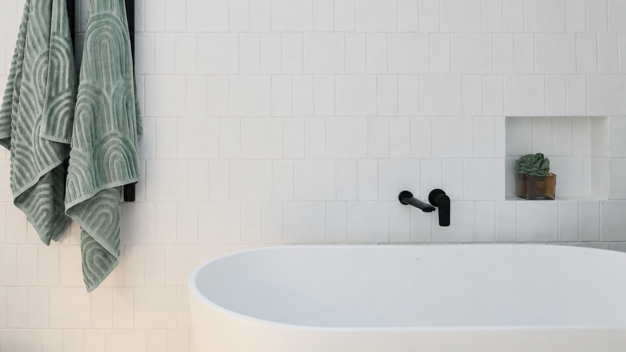 A minimalist bathroom with a white freestanding bathtub, black wall-mounted faucet, a small tile wall niche with a green towel, and two green towels hanging on a rack on the left side.