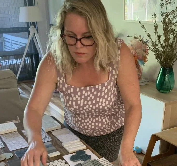 Interior designer with blonde wavy hair and glasses standing over a table with materials samples with a window and green vase with flowers in the background.