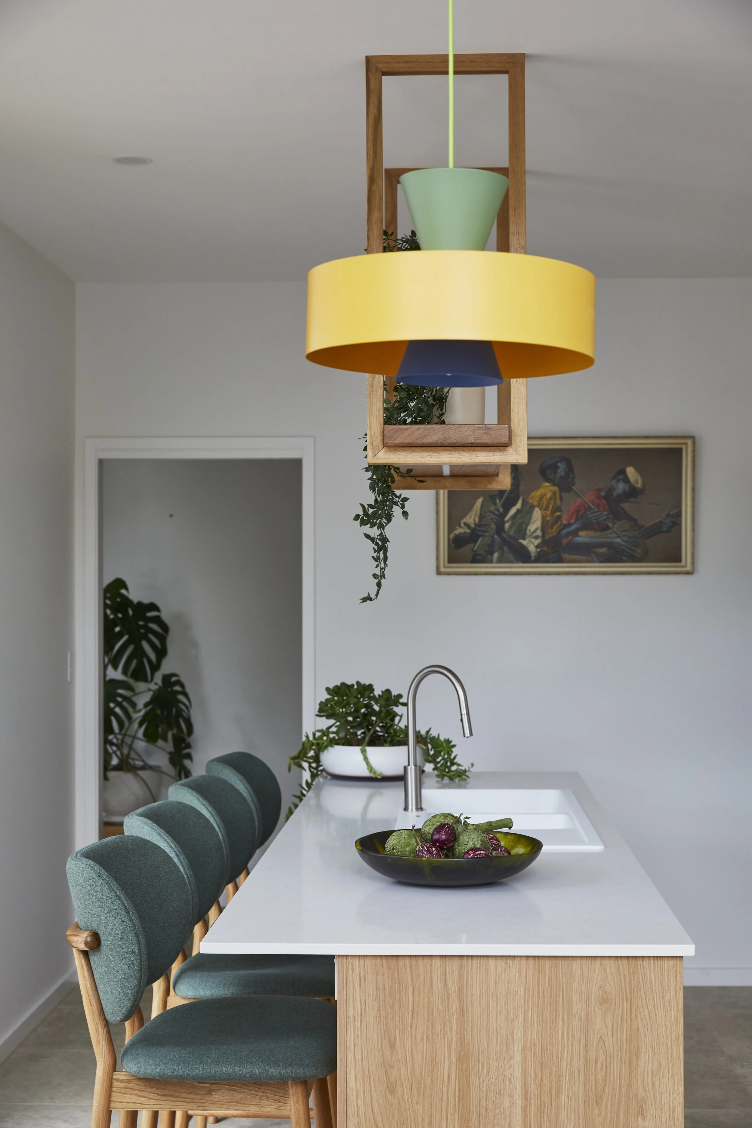 Modern kitchen with a white island counter, green cushioned chairs, and a black bowl filled with vegetables, including purple onions. Above the island, a colorful pendant light with a yellow, green, and blue shade hangs. In the background, there's a framed painting of jazz musicians, and some plants are visible near a doorway.