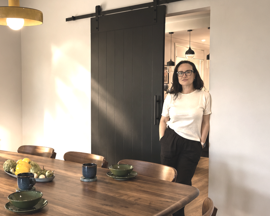 Content client in glasses wearing a white t-shirt and black pants standing in doorway of modern dining room with a wooden table, bowls, and mugs.