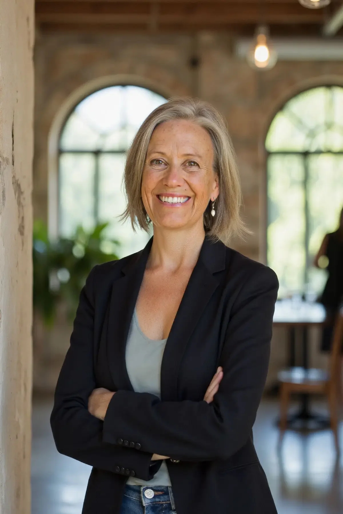 Pip Smith Health, wearing a black blazer and earrings, standing with arms crossed in an industrial-style office with large windows and brick walls.