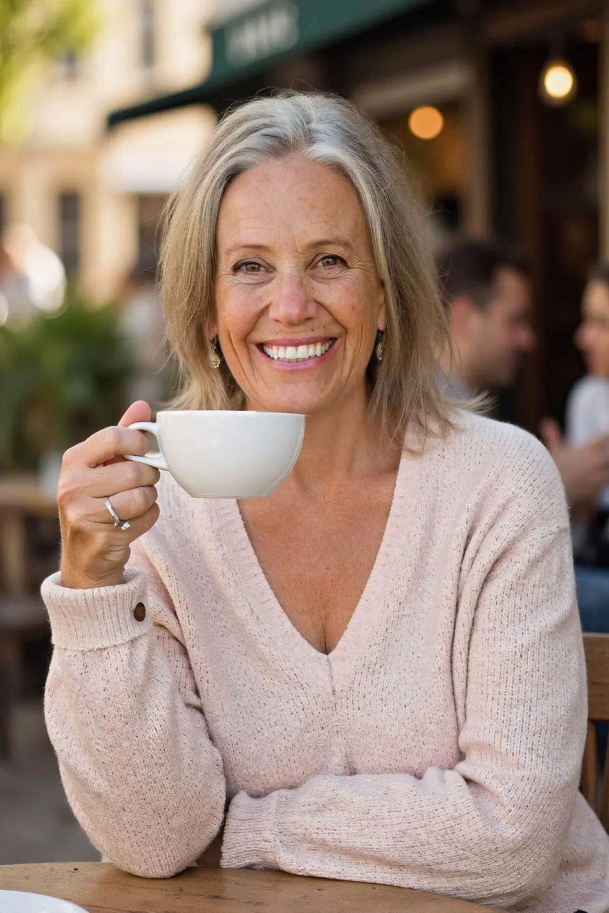 A smiling older woman with gray hair holding a white coffee cup at an outdoor café.