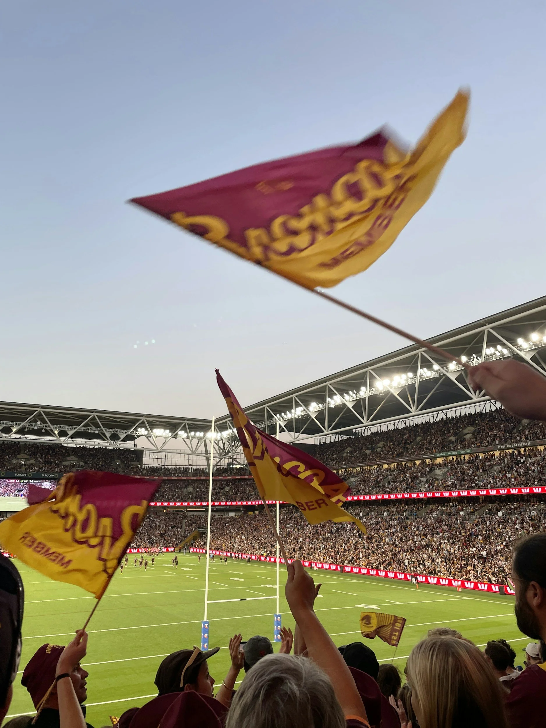Fans waving Brisbane Bronchos maroon and yellow flags at a football stadium filled with spectators during a game, with players visible on the field and the stadium's roof and bright lights overhead.