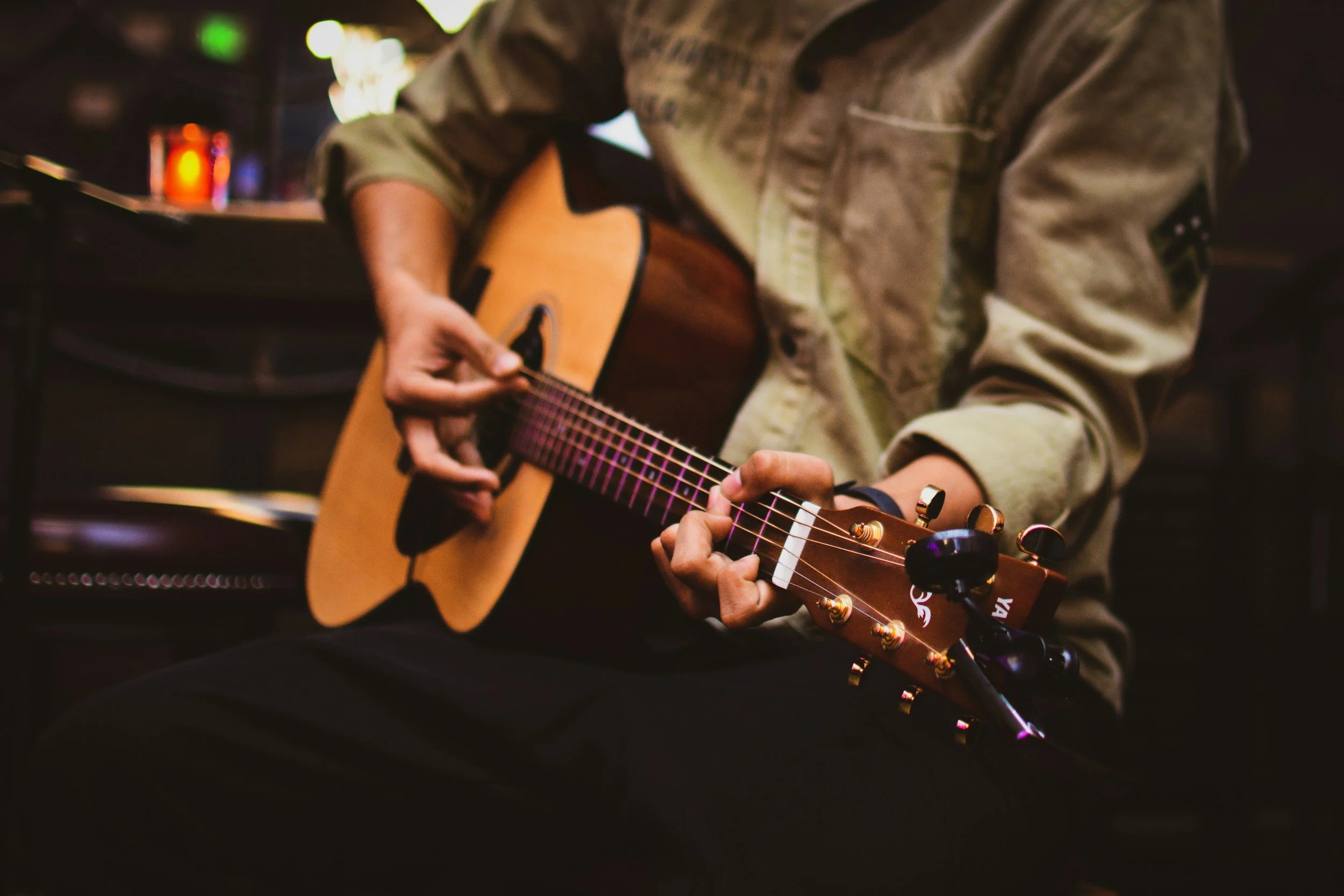 Person wearing a green jacket playing an acoustic guitar in a dimly lit room.
