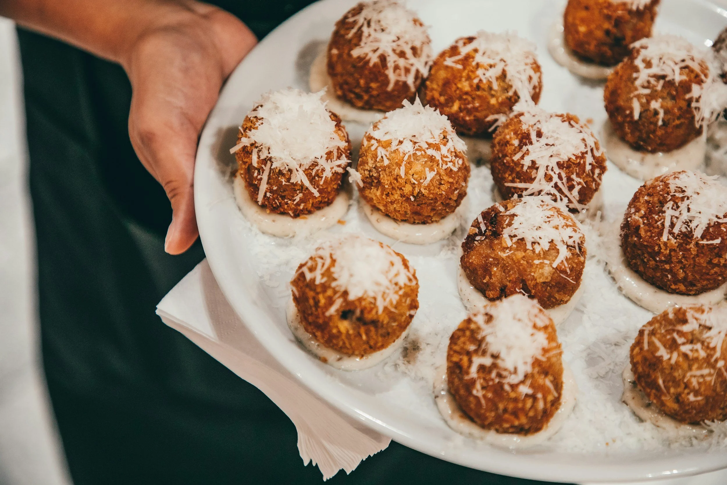 A white plate of fried croquettes topped with shredded cheese, resting on a napkin.