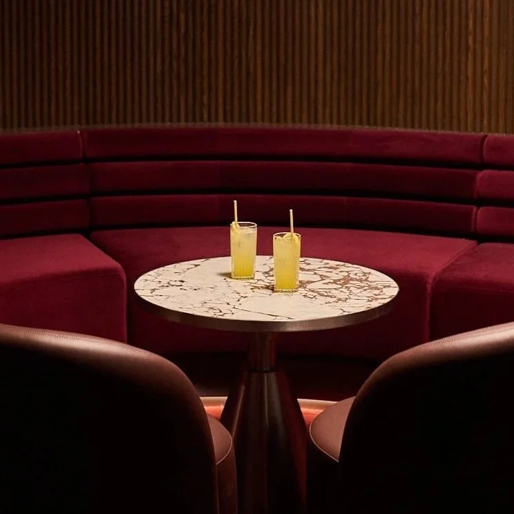 Two yellow drinks with straws on a white marble round table surrounded by red upholstered seating in a dimly lit Vixen Room