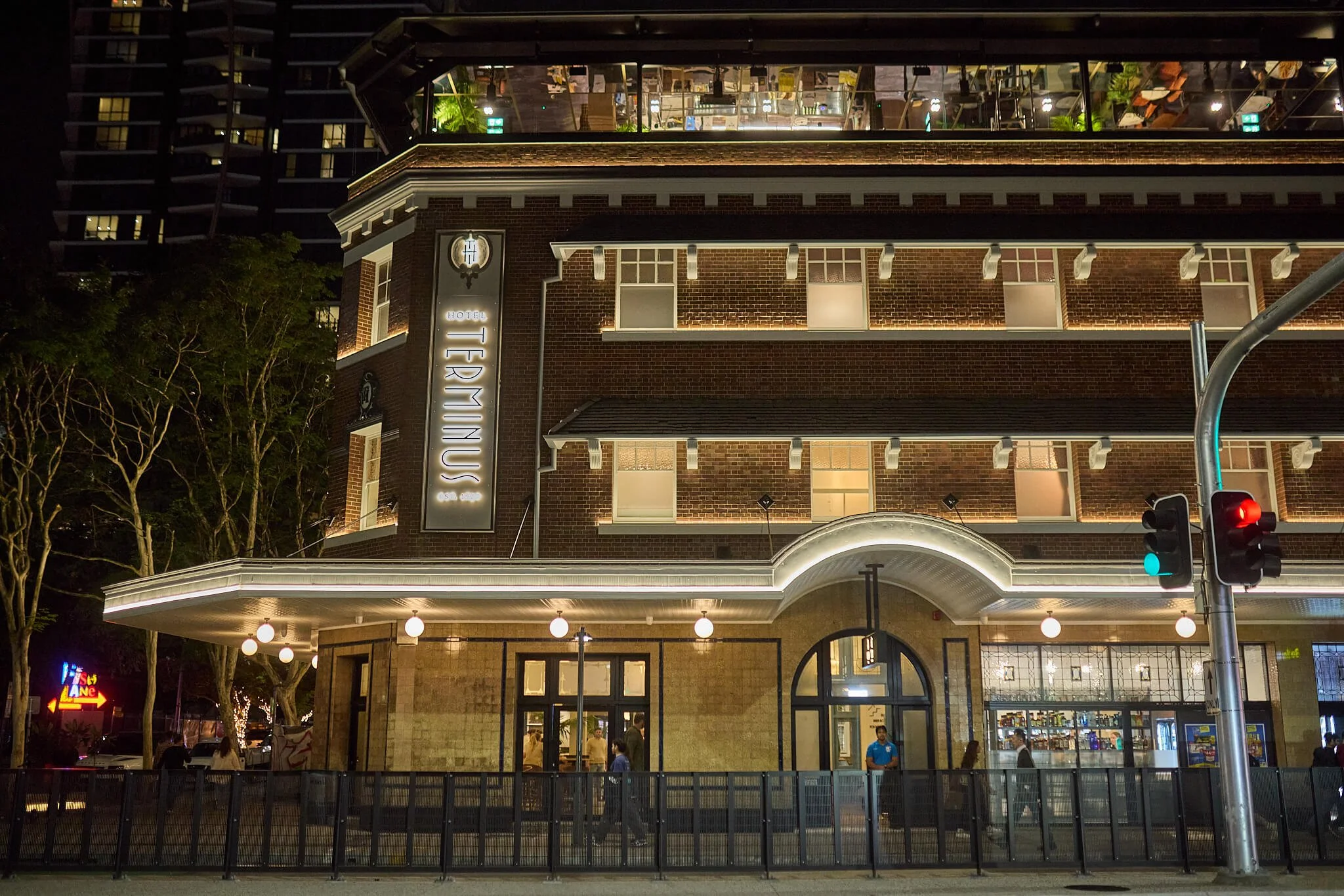 Nighttime view of a multi-story Hotel Terminus with illuminated sign, outdoor lighting, and visible interior spaces. People are gathered outside.