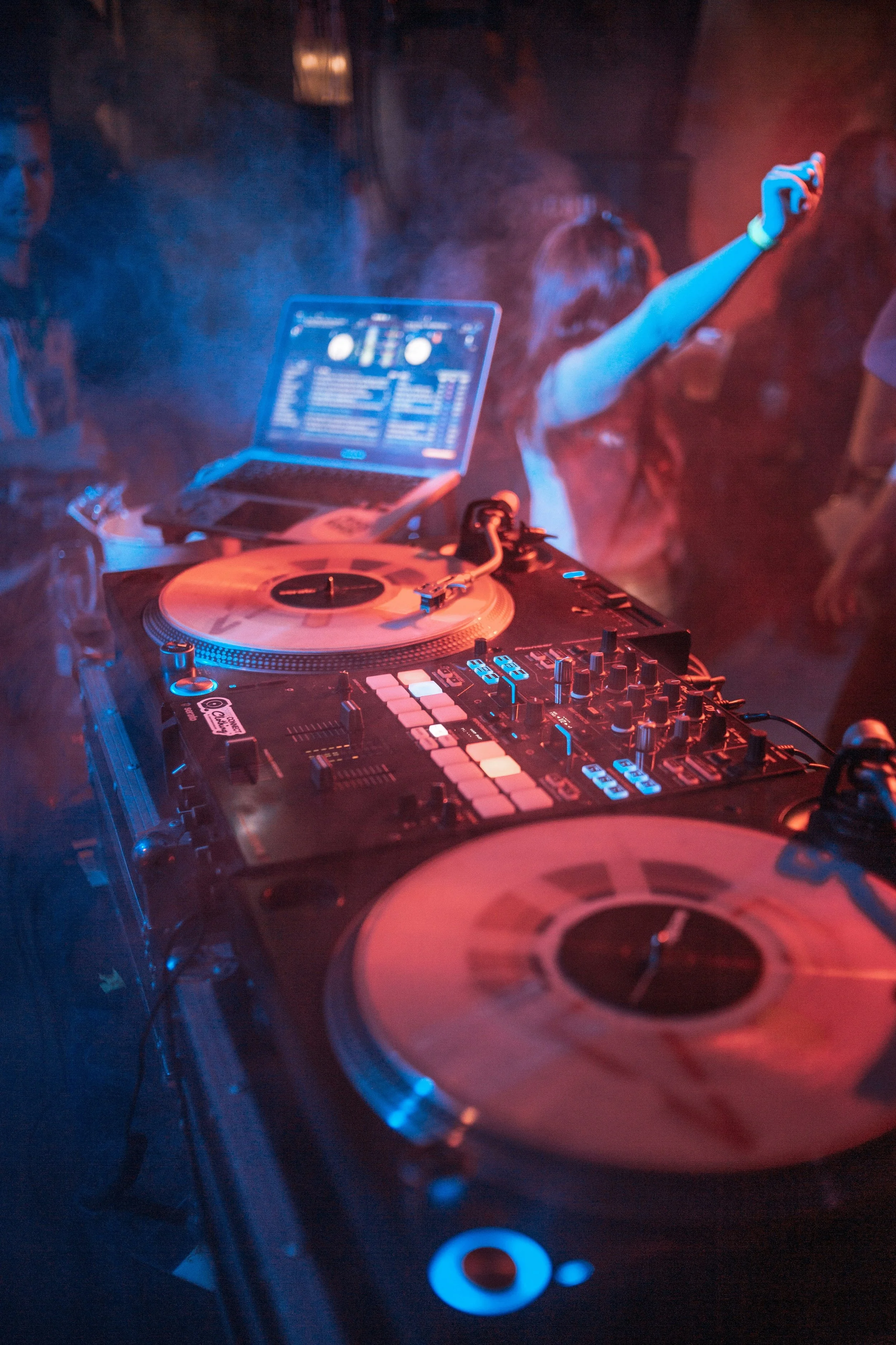A DJ setup with turntables and a laptop at a party or club, with people dancing in the background.