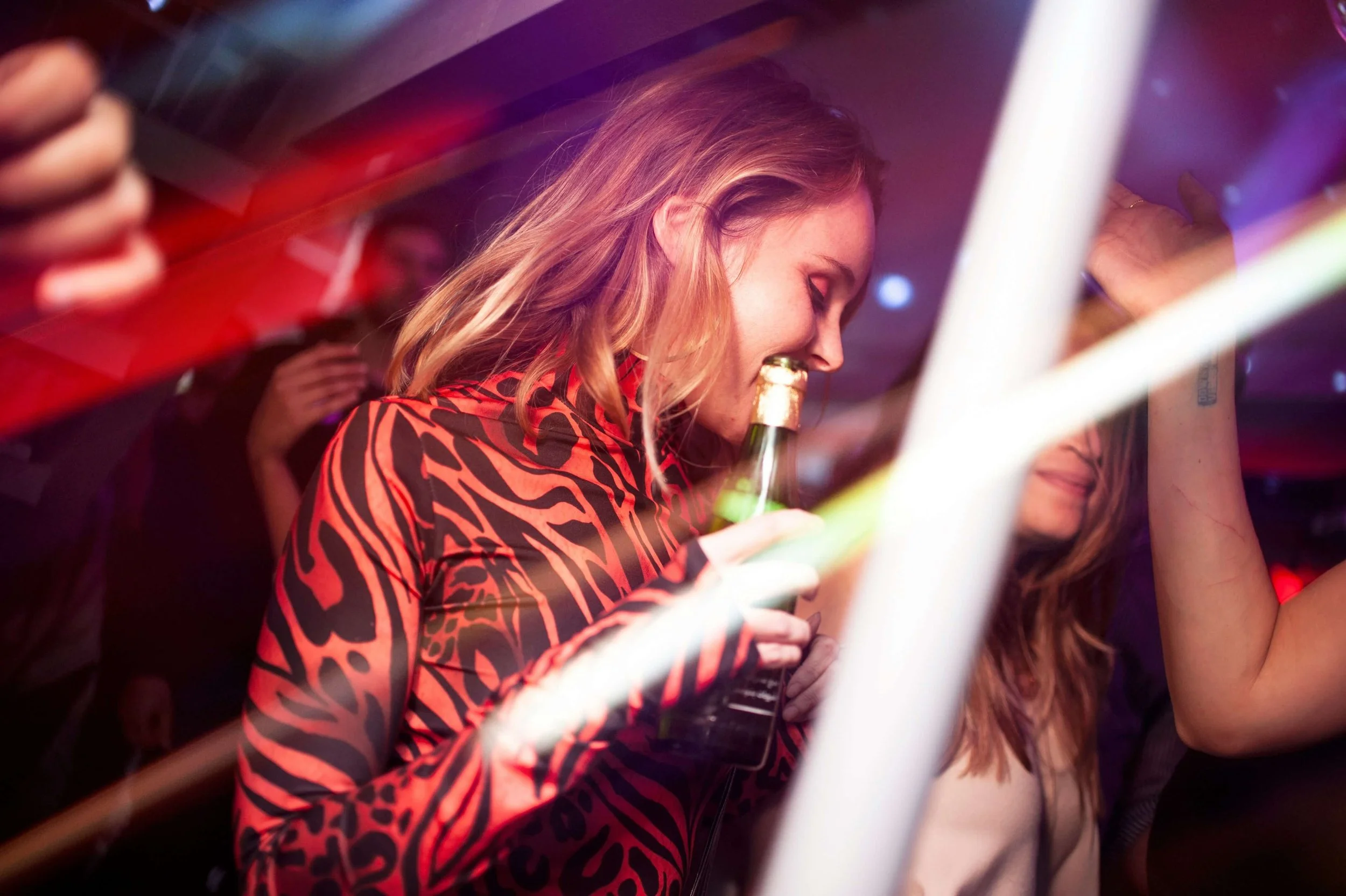 A woman with light skin and wavy blonde hair, wearing a red and black patterned dress, smiling and holding a beer bottle at a party or nightclub.