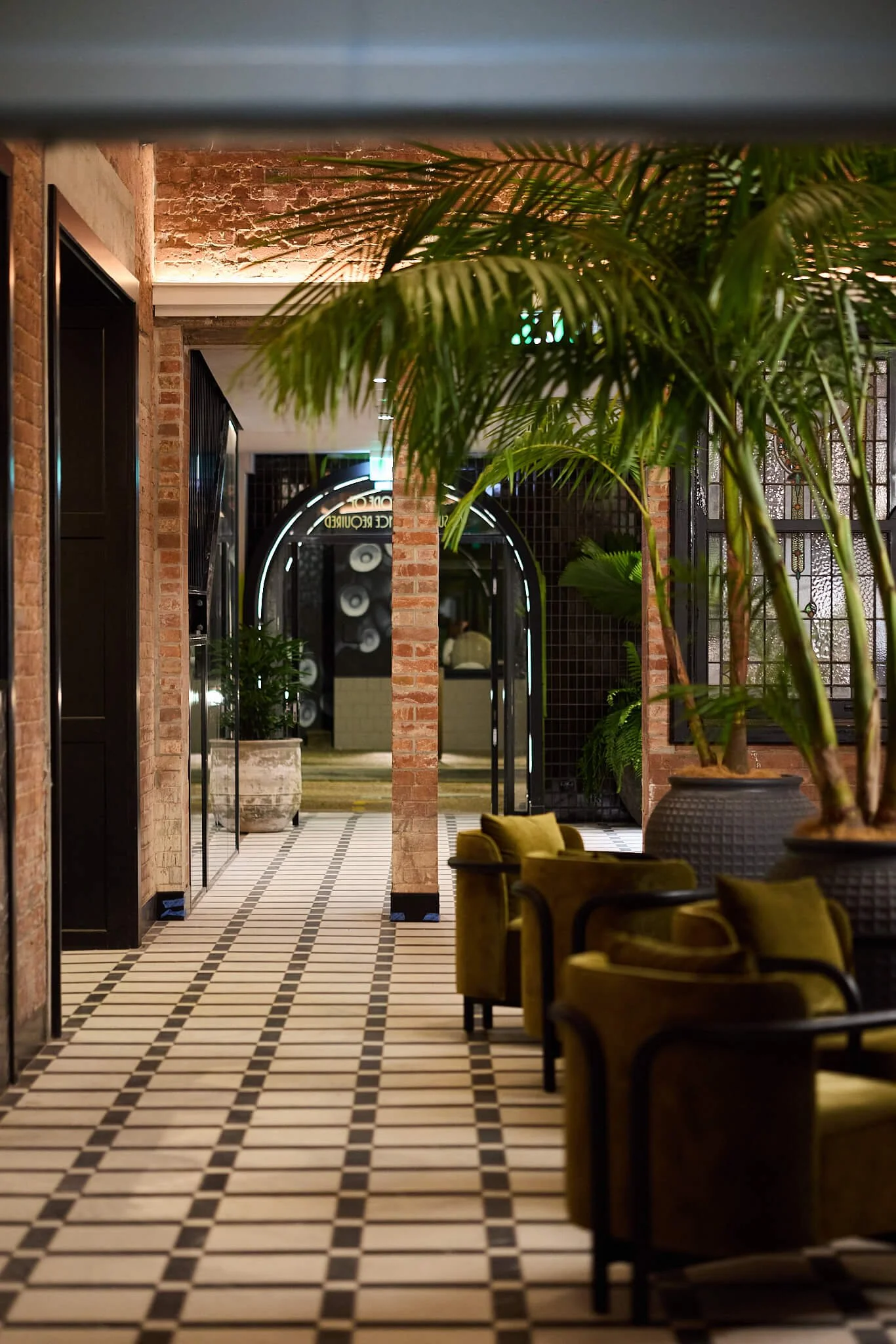 Interior of a Hotel Terminus with black and white checkered flooring, exposed brick walls, large potted plants, and green velvet chairs.