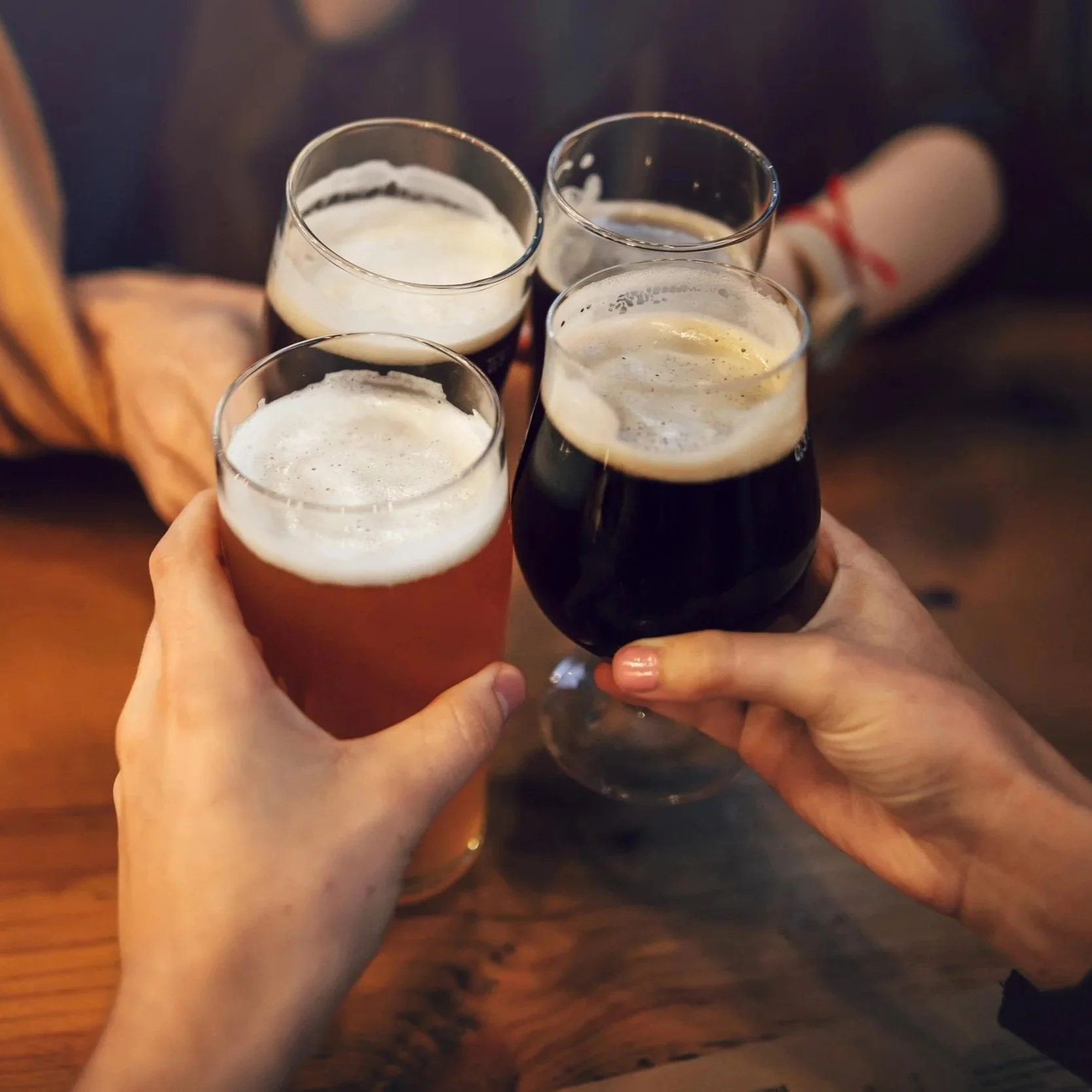 Four hands clinking glasses of beer in a toast over a wooden table.
