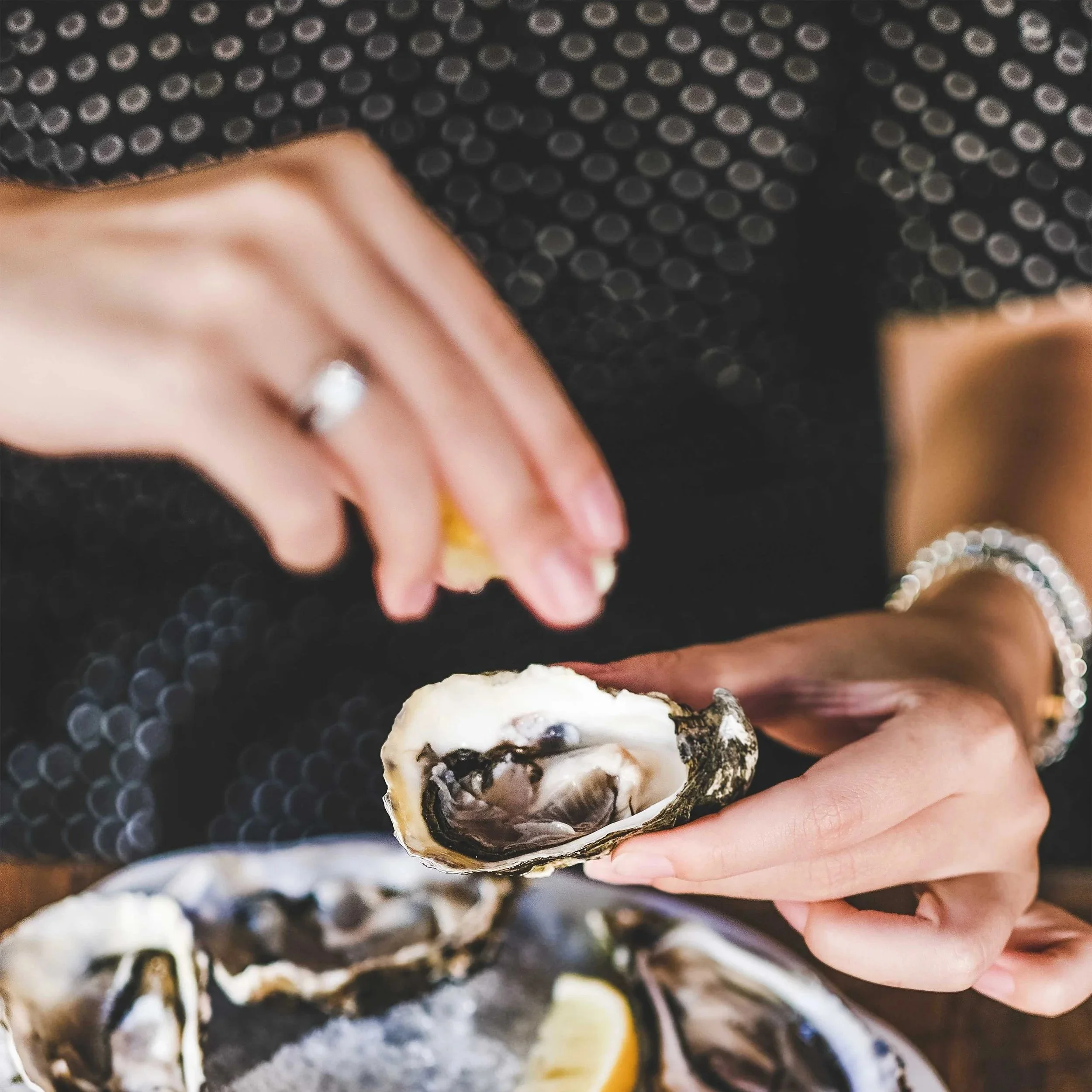 Close-up of a hand holding an oyster shell with pearls inside, against a dark, dotted background.