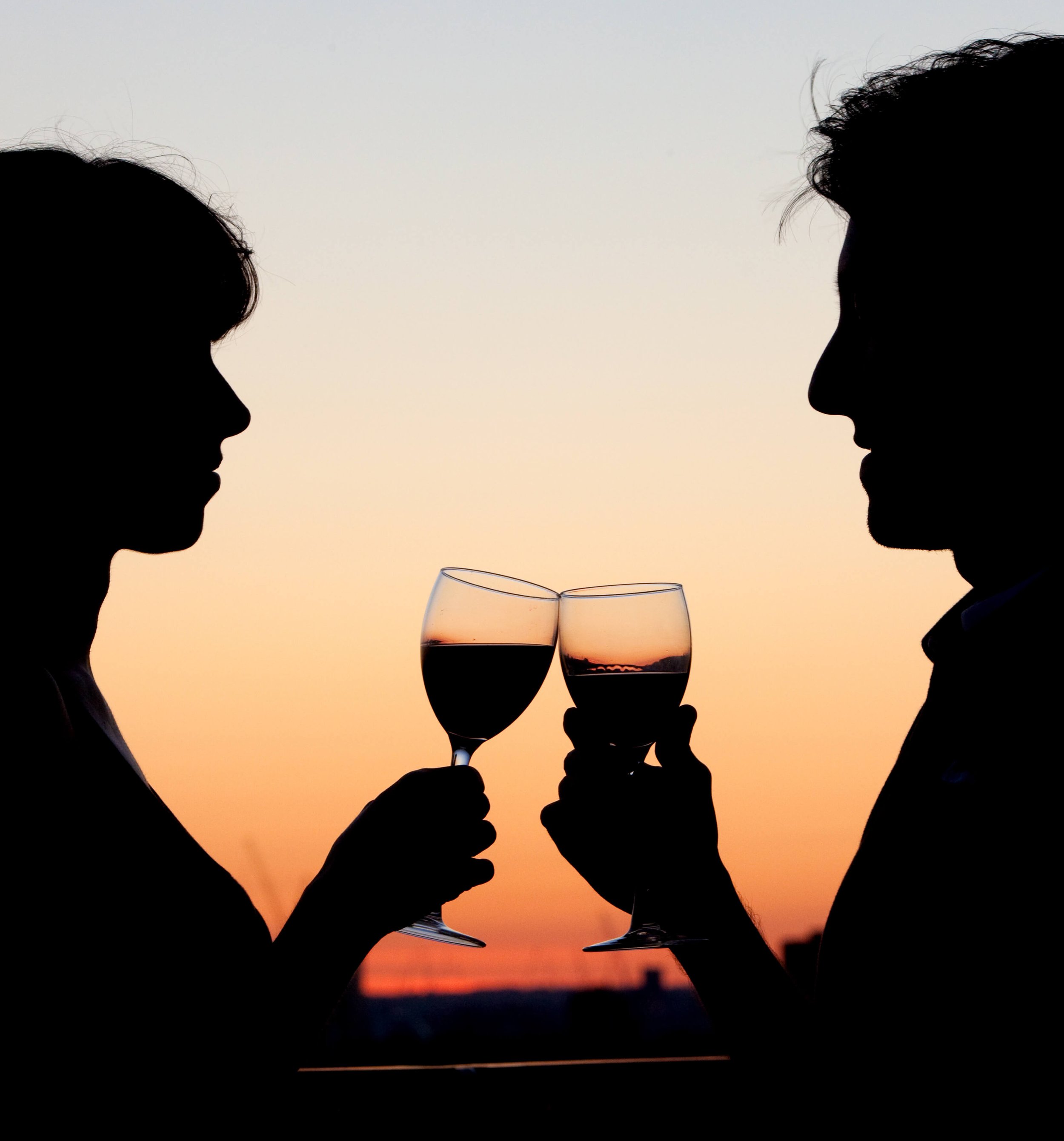 Silhouettes of a man and a woman holding glasses of red wine facing each other during sunset.