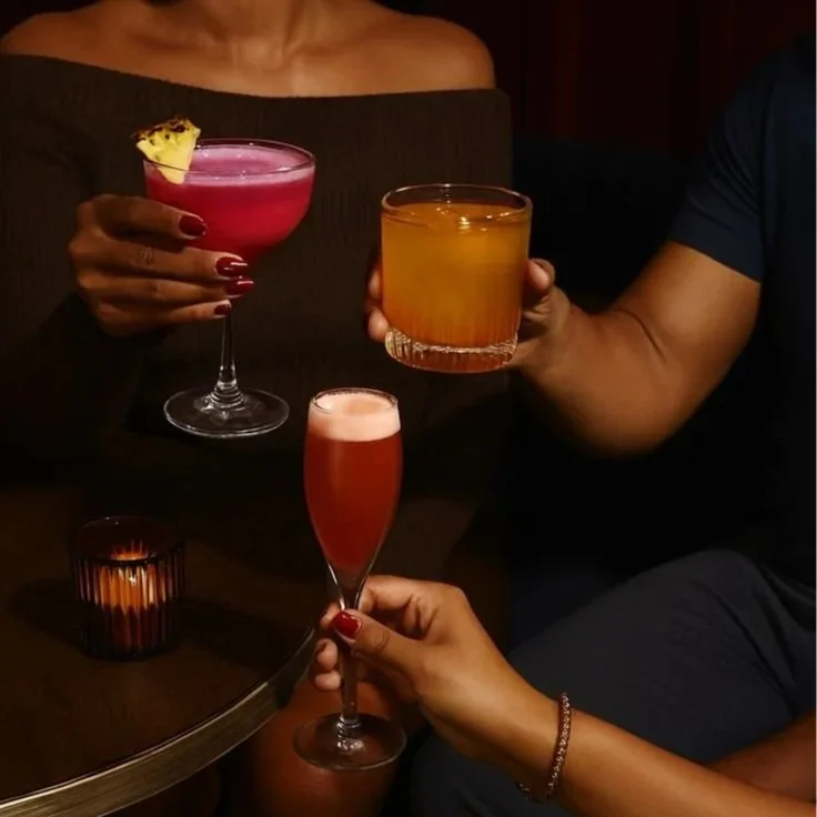 Three people holding colorful cocktails or drinks in a dimly lit setting, with a small candle on the table.