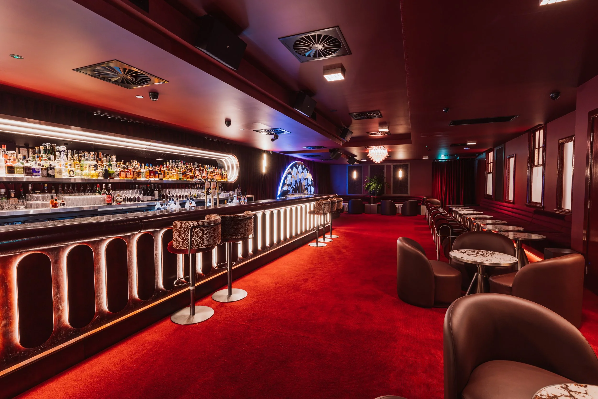 Dimly lit indoor bar with a curved bar counter displaying various liquor bottles on shelves, surrounded by barstools and small round tables with lamps, and dark red curtains in the background.