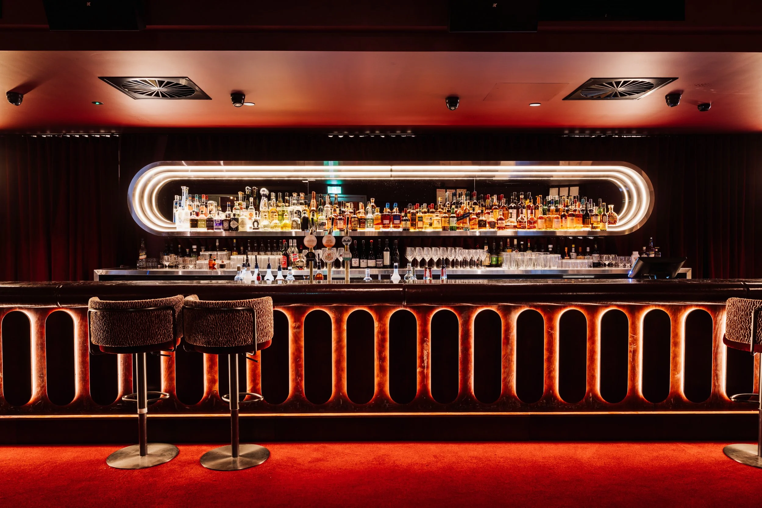 A sophisticated bar with a neon fox logo and the name 'Vixen Room' on the glass window, featuring a dark ambiance, wooden bar counter, bar stools, and small tables with lamps.