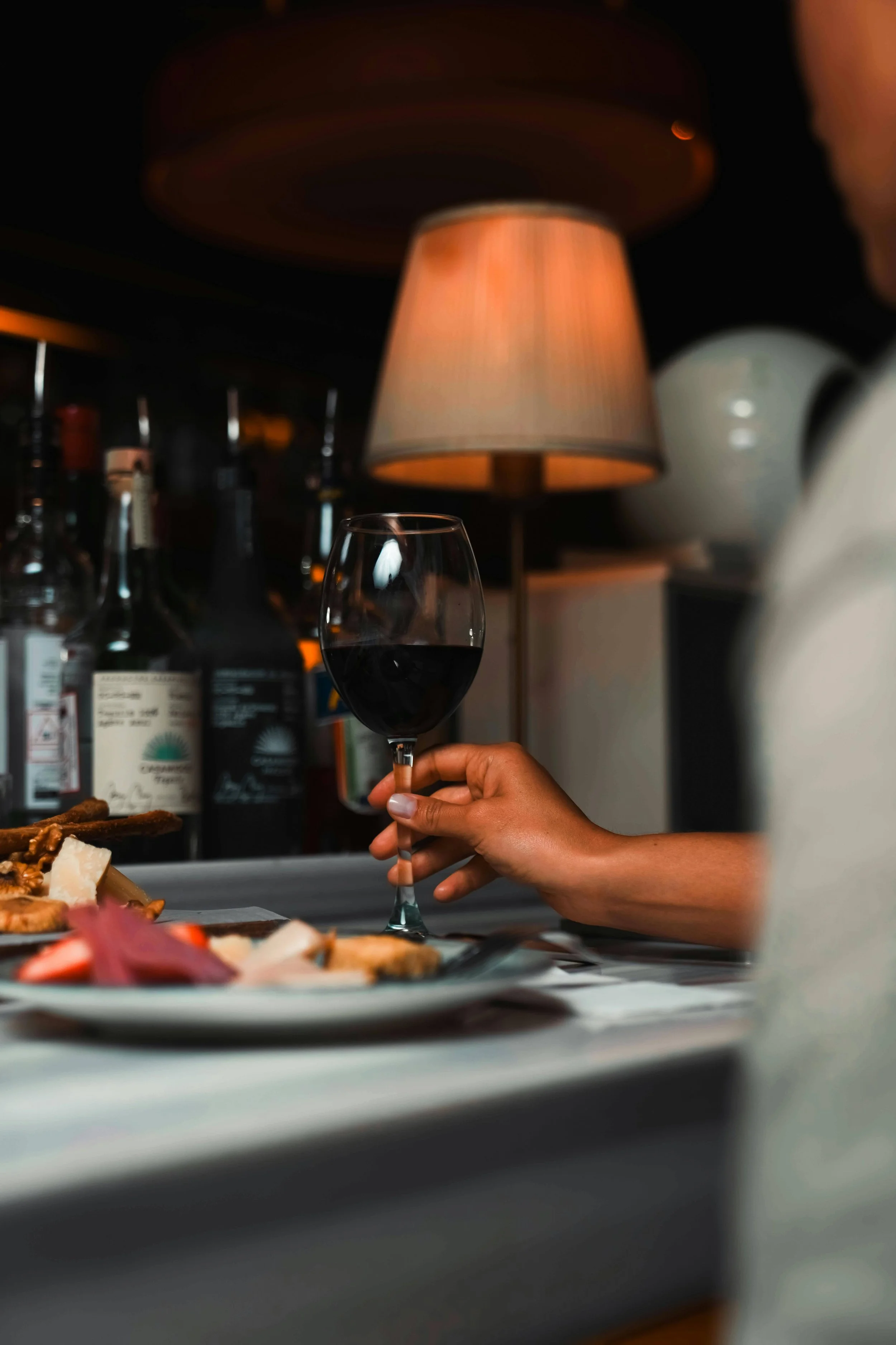 A person holding a glass of red wine at a dinner table, with a blurred background of bottles and a warm, orange lamp.