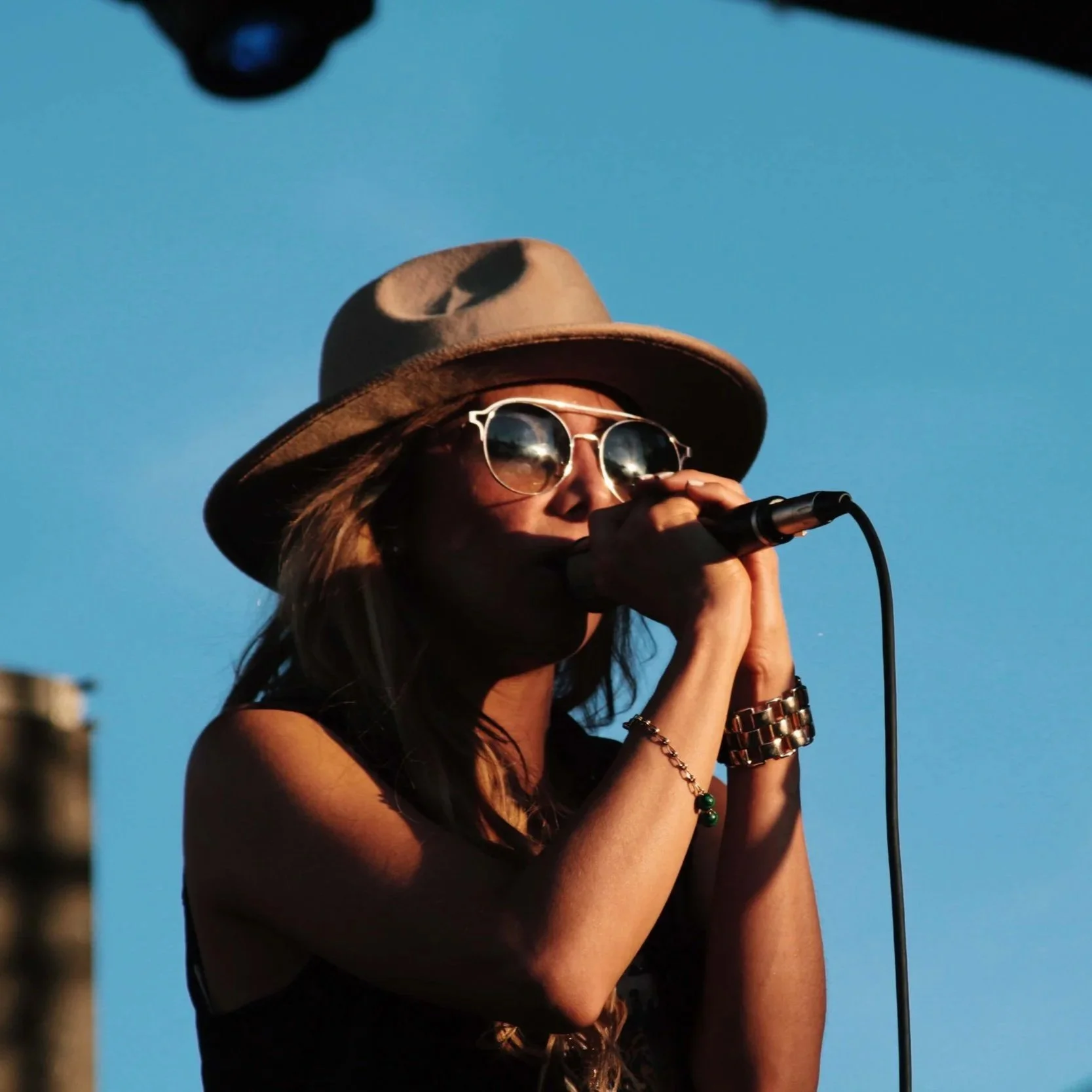 A woman wearing a wide-brimmed hat, sunglasses, and jewelry, singing into a microphone outdoors against a clear blue sky.