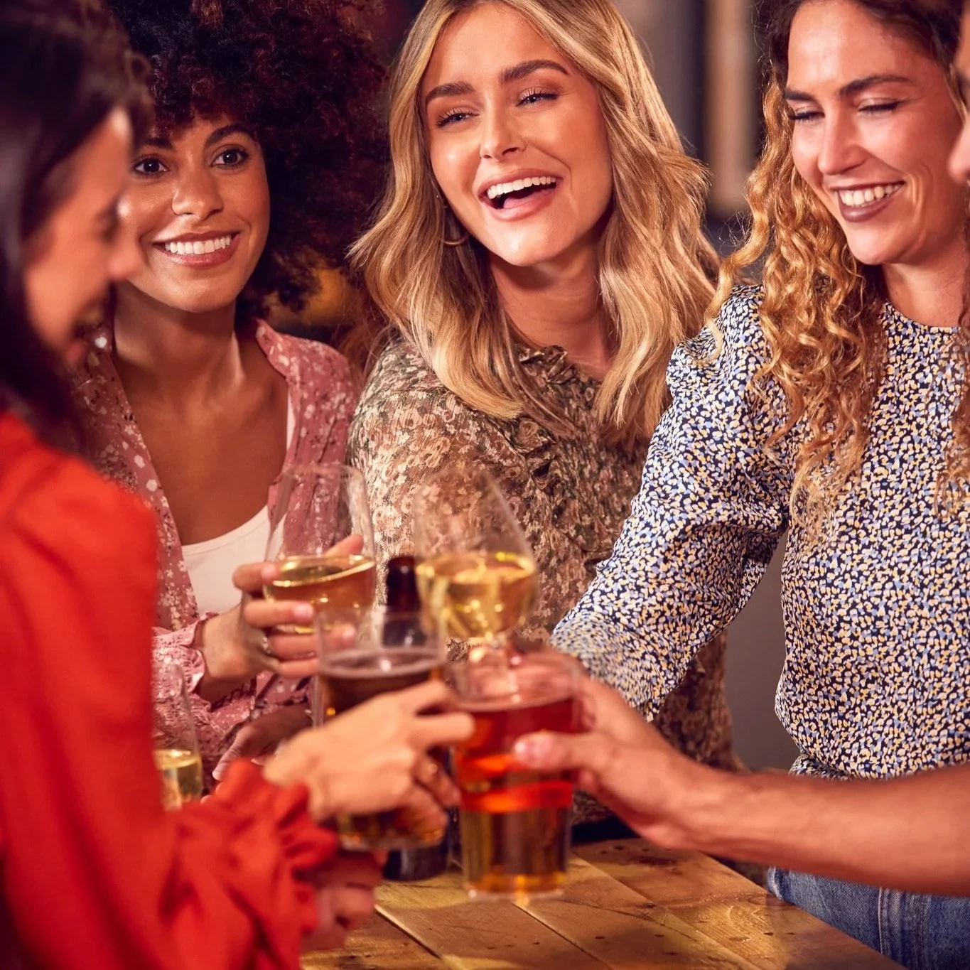 Group of women smiling and clinking glasses of wine and beer in a social setting.