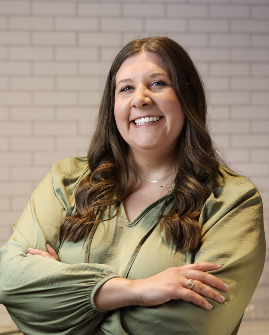 Headshot of founder, Elizabeth Pearce, smiling with arms crossed, wearing a sage green blouse and cross necklace, standing in front of a softly blurred white brick background.