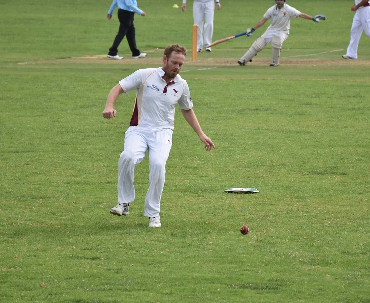 A man dressed in white cricket uniform is about to catch a cricket ball on a grassy field, with other players in white in the background.