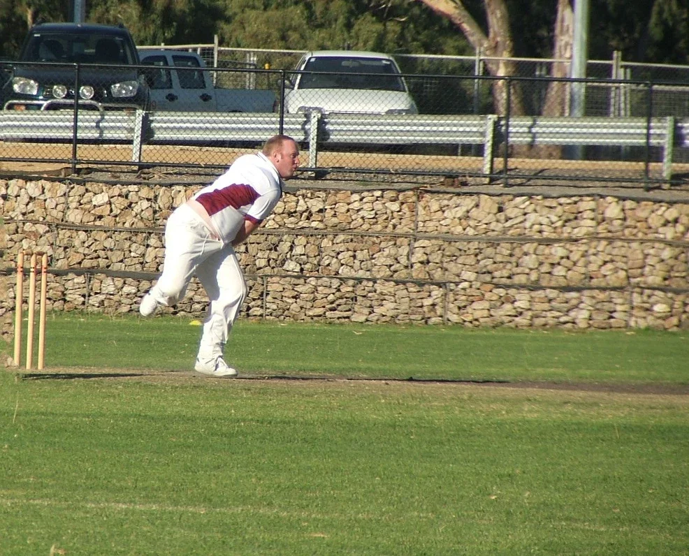 A man in cricket attire bowing on a cricket field next to the stumps, with cars and a stone wall in the background.