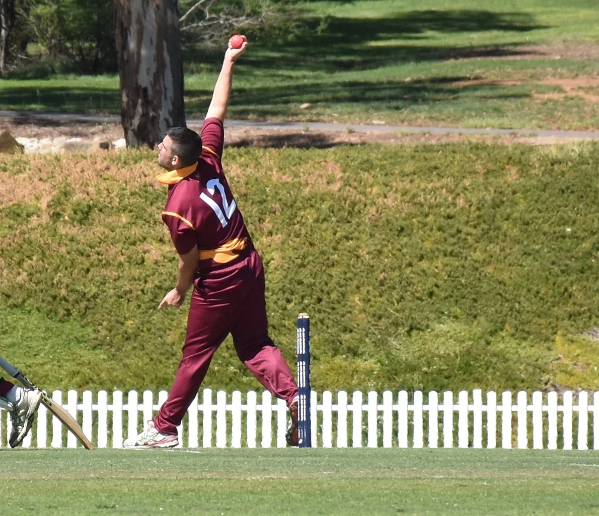 A cricket player in a maroon and yellow uniform bowling on a cricket field.
