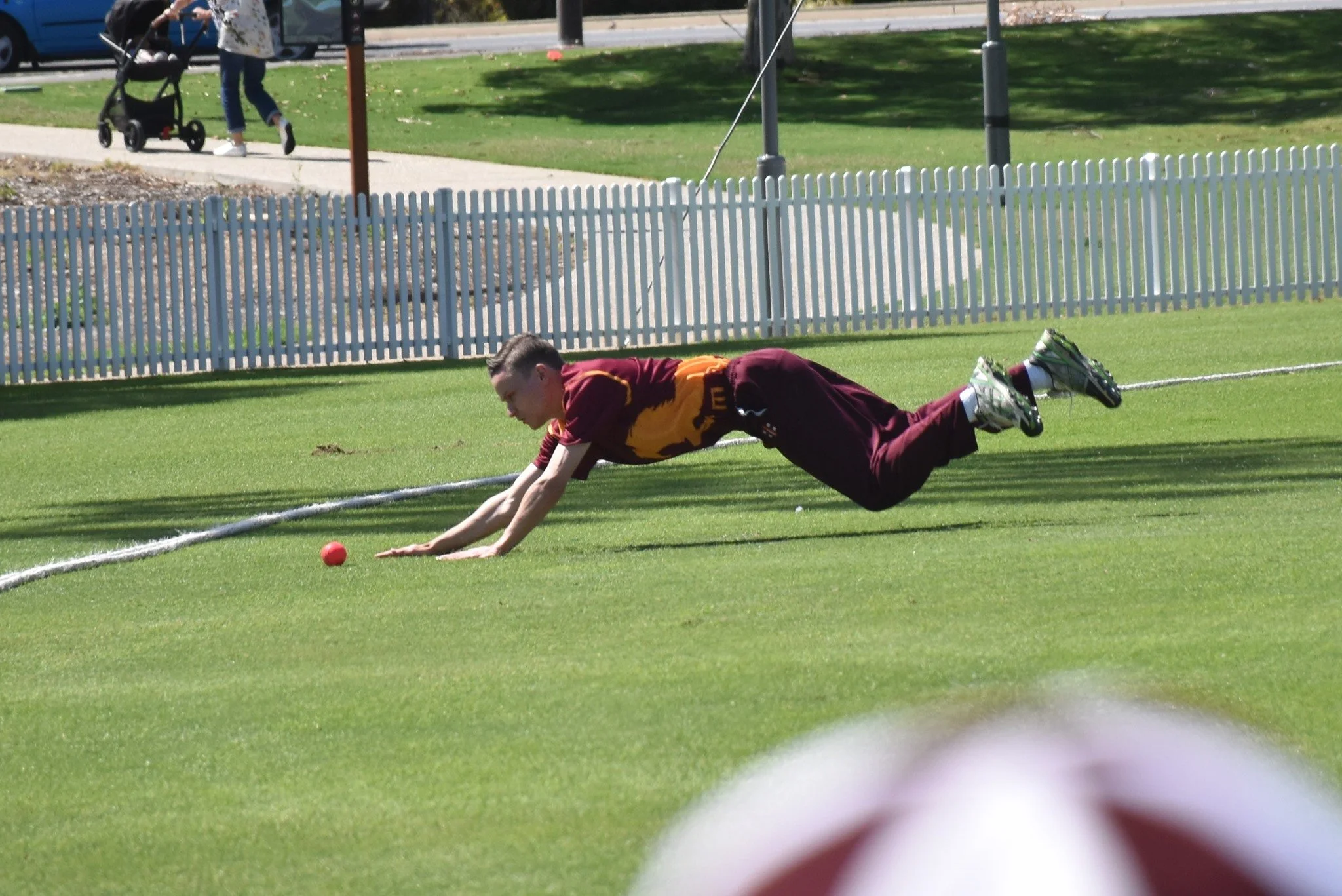 A person dressed in sportswear is diving on a grassy sports field to retrieve a small red hockey ball, with a white boundary line in the foreground and a white picket fence in the background.