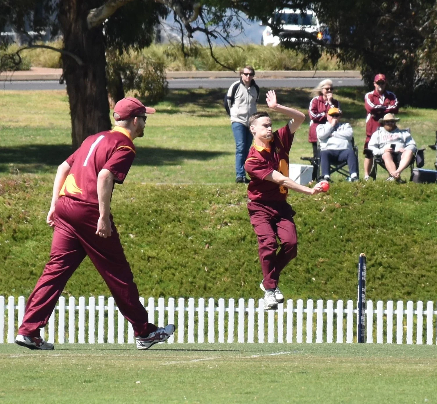 A cricket player in maroon uniform jumping to hit the ball while another person in maroon is walking nearby. Spectators are sitting and standing in the background near trees.