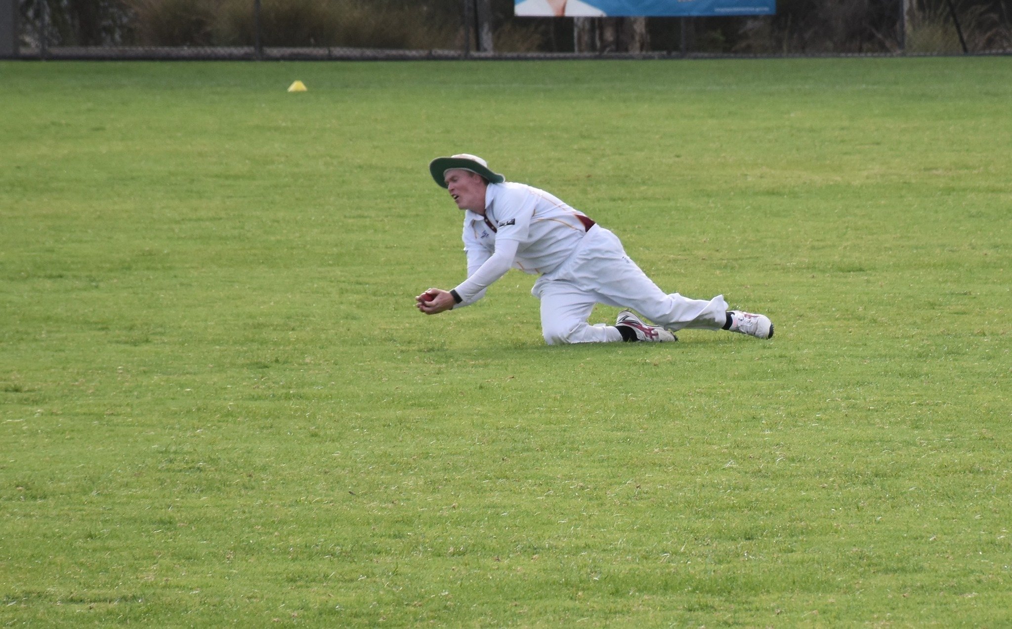 A person wearing a white cricket uniform and a wide-brimmed hat is kneeling on the grass field, holding a cricket ball with both hands, appearing to be fielding or preparing to throw.