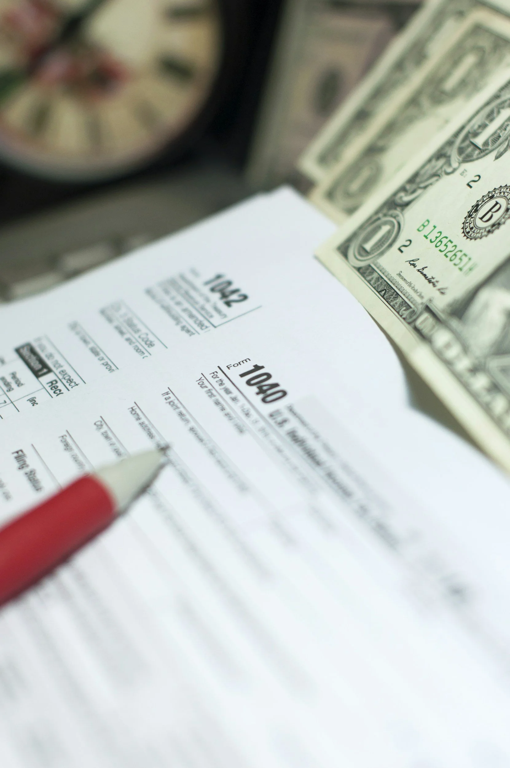 A close-up of a tax form (1040) on a desk, with a red pen, a clock, and a folded stack of U.S. dollar bills in the background.