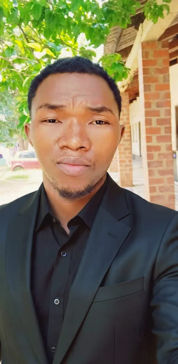 A young man in a black suit and shirt taking a selfie outdoors, with green leafy trees and a brick building in the background.