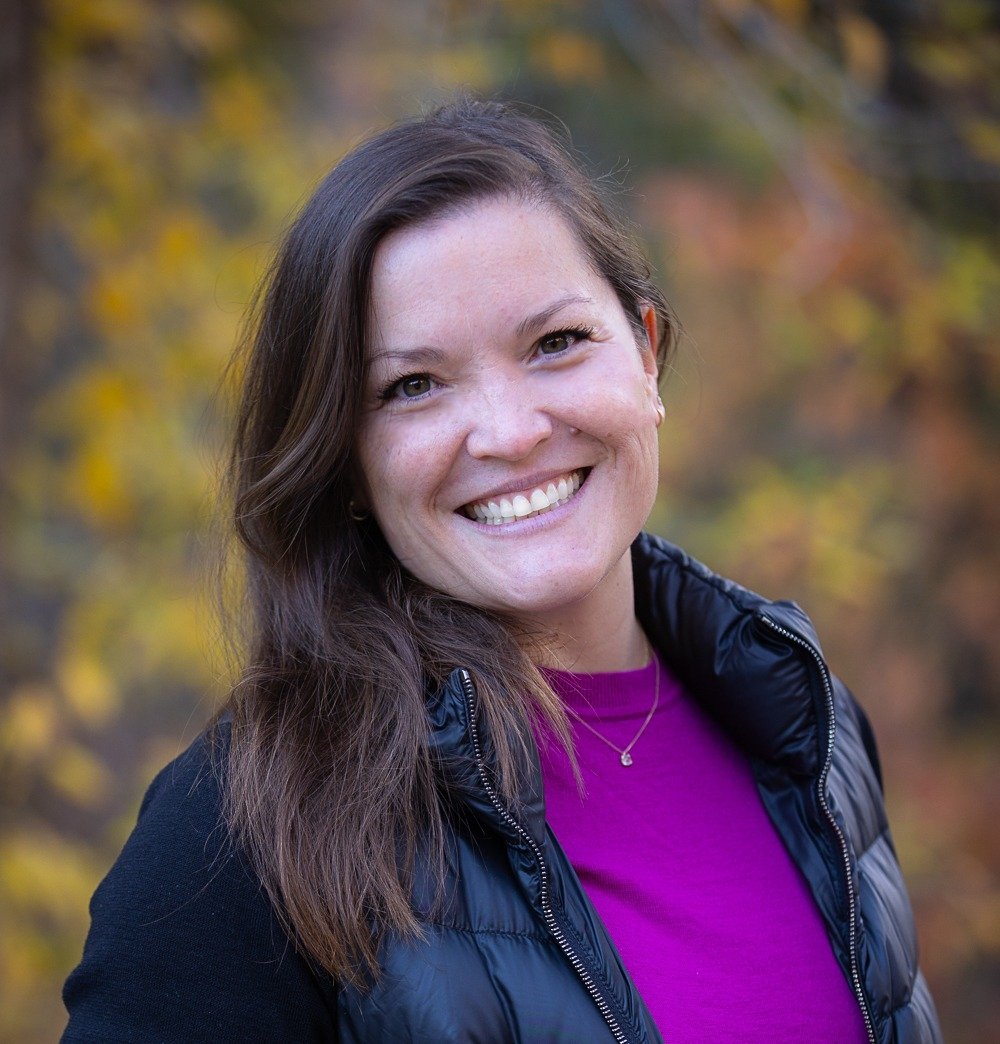 Smiling woman with long brown hair, wearing a purple shirt and a black puffer jacket outdoors with autumn foliage in the background.