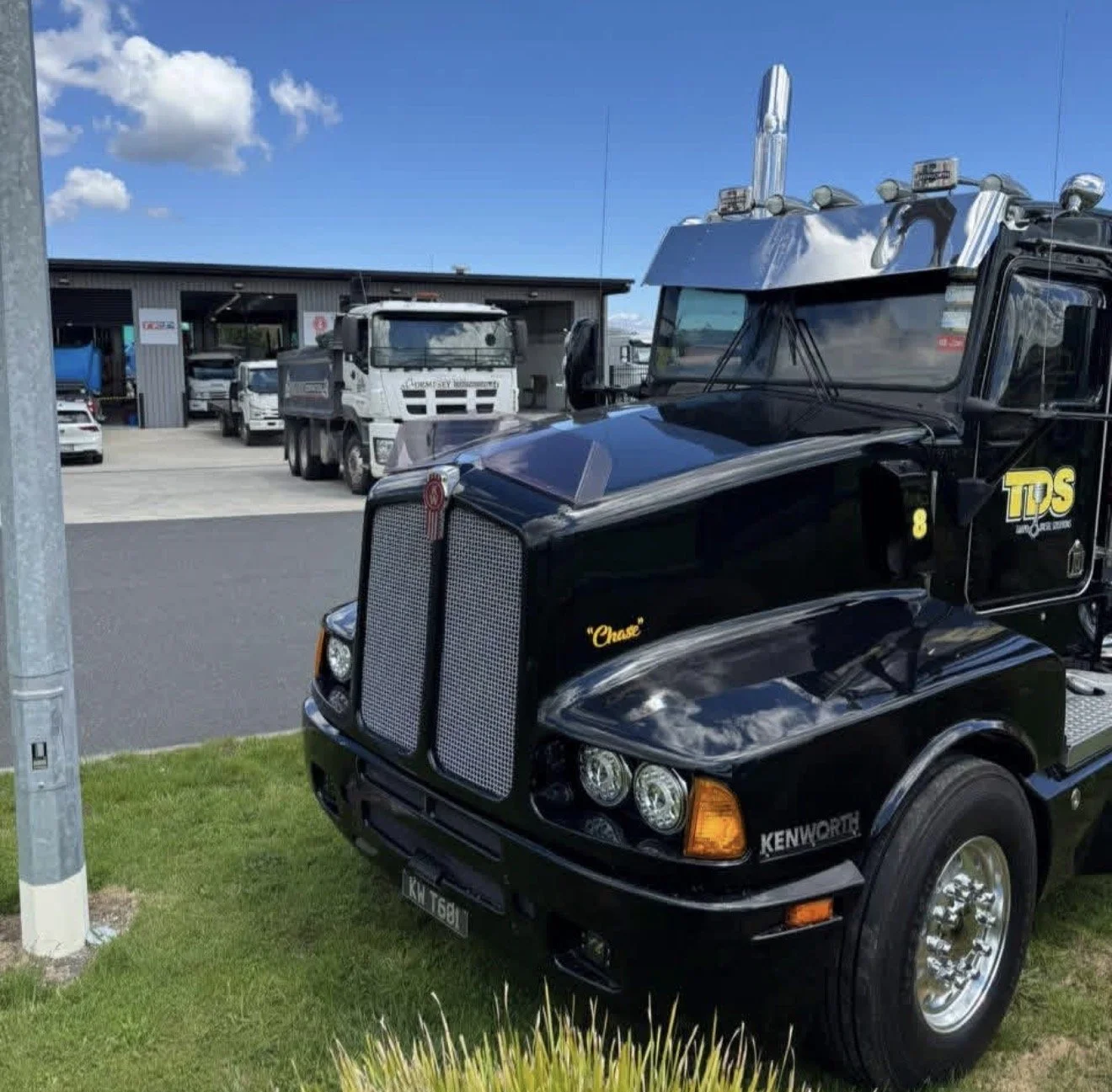 A black semi-truck parked in front of a service or repair building, with several other trucks and cars in the background on a sunny day with a blue sky and some clouds.