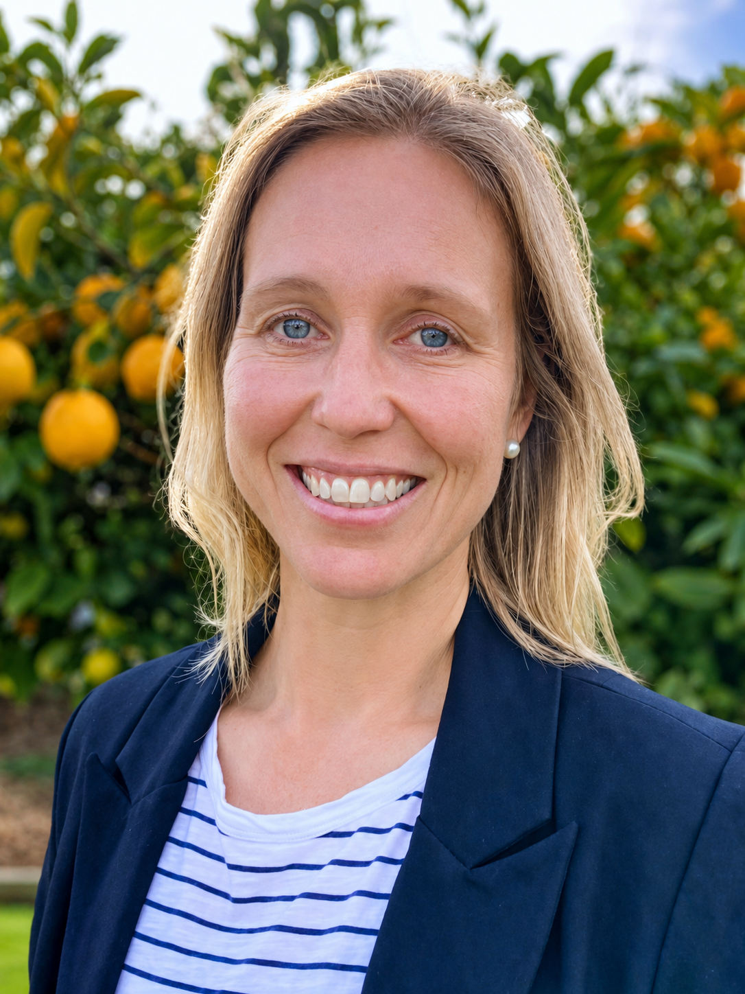 A woman with blonde hair, blue eyes, and a bright smile wearing a navy blazer and pearl earrings, standing in front of a citrus tree with ripe yellow oranges.