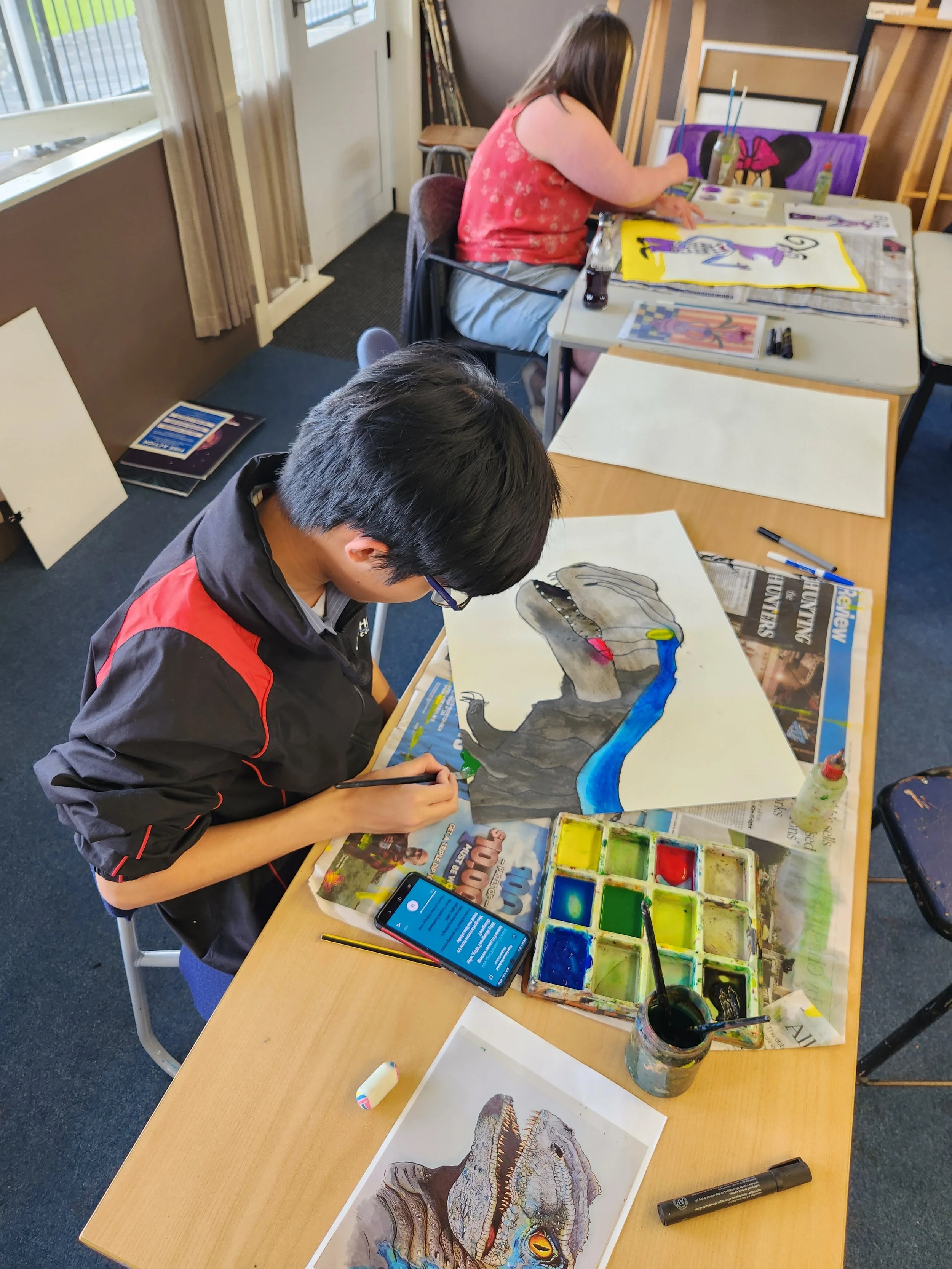 Two children sitting at a table working on dinosaur-themed art projects. The girl in a red shirt paints with watercolors, and the boy in a black and red jacket sketches a dinosaur illustration. Art supplies and colored water paints are scattered across the table.