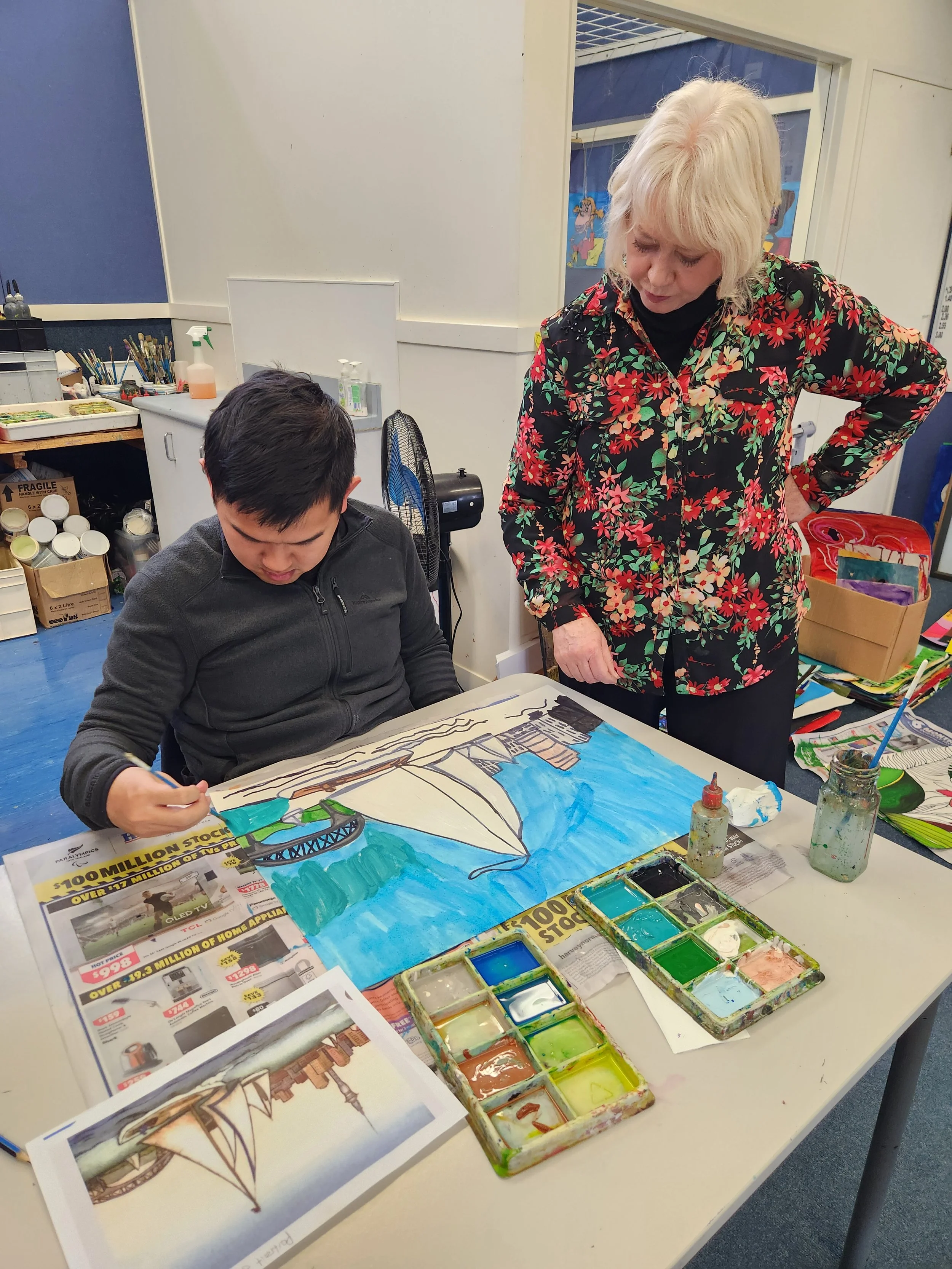 A child and an adult are working on an art project at a table. The child is painting a large poster depicting a bridge and water scene with trees. The table has watercolor paints, brushes, and printed reference images, with newspapers laid out underneath.