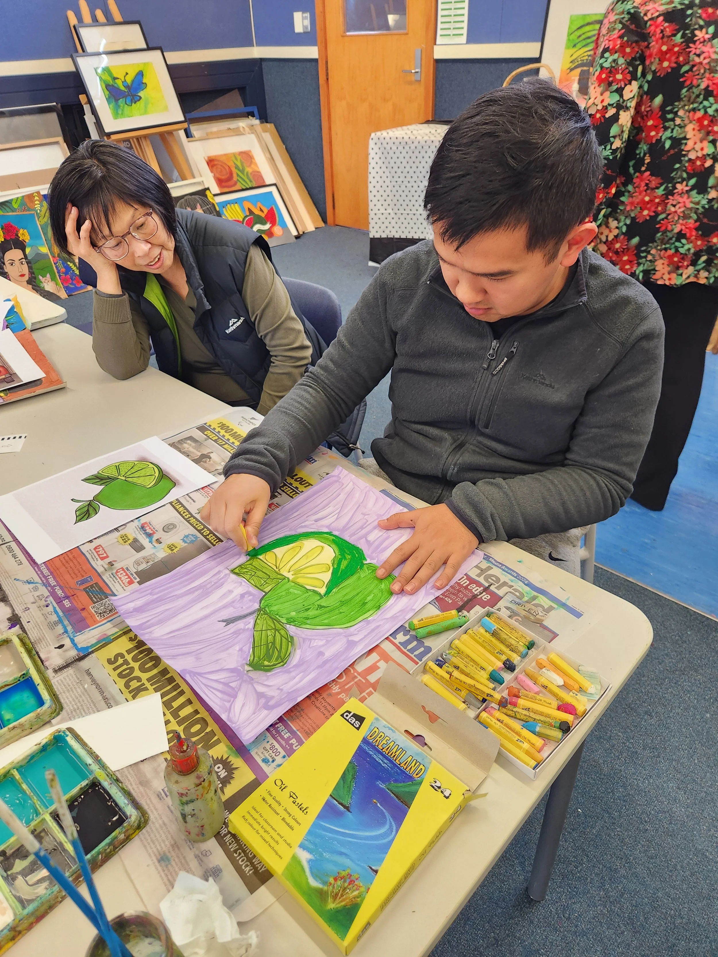 Two people sit at a table covered with art supplies and media, working on a colorful painting of a large green lime with yellow segments. The art supplies include pastel chalks, paints, brushes, and printed images of similar artworks. In the background, framed paintings of various colorful artworks are displayed against a blue wall with a wooden door.