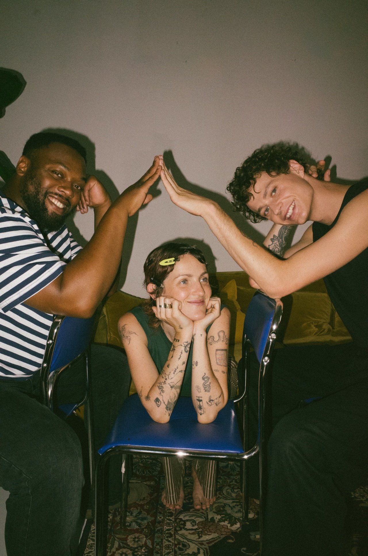Three people are sitting in a room with a patterned rug. Two people are standing and engaging in a friendly high-five, one man with a beard wearing a striped shirt and a woman with curly hair and tattoos. The third person, a woman with short hair and numerous tattoos, is seated between them, smiling with her hands resting on her chin, watching the interaction.