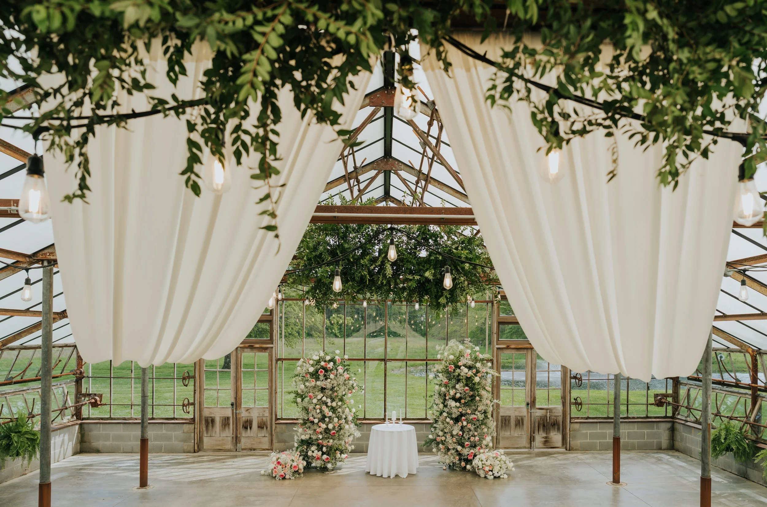 A rustic greenhouse wedding setup with white curtains, hanging light bulbs, and floral arrangements of pink and white flowers, with greenery sprigs hanging from the ceiling.