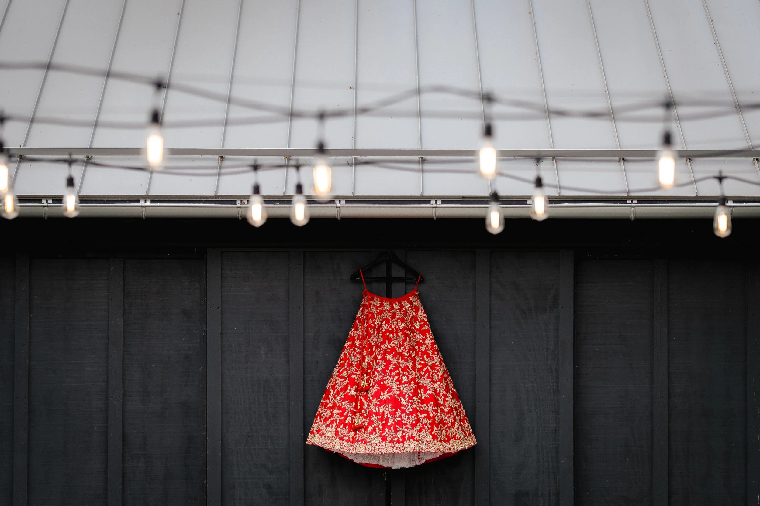 A red wedding dress with gold floral embroidery hanging on a black wall beneath a modern ceiling with exposed bulbs and a gray metal roof.