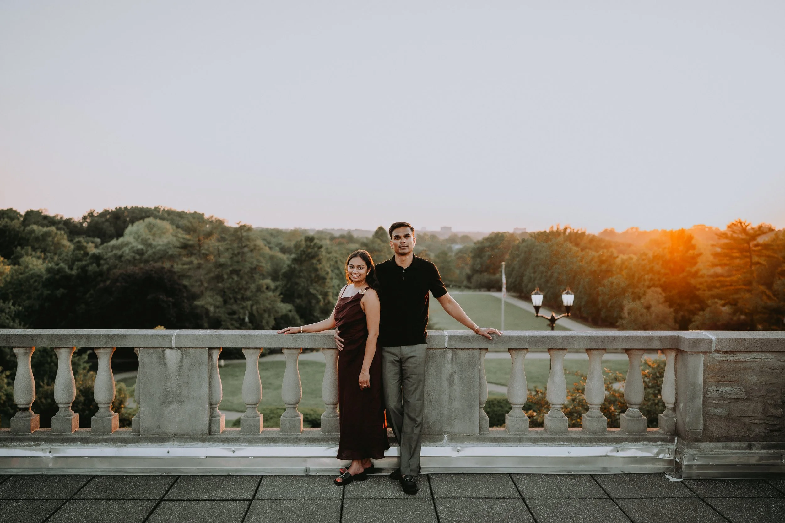 A young couple standing on a balcony at sunset with a scenic view of trees and a park in the background.