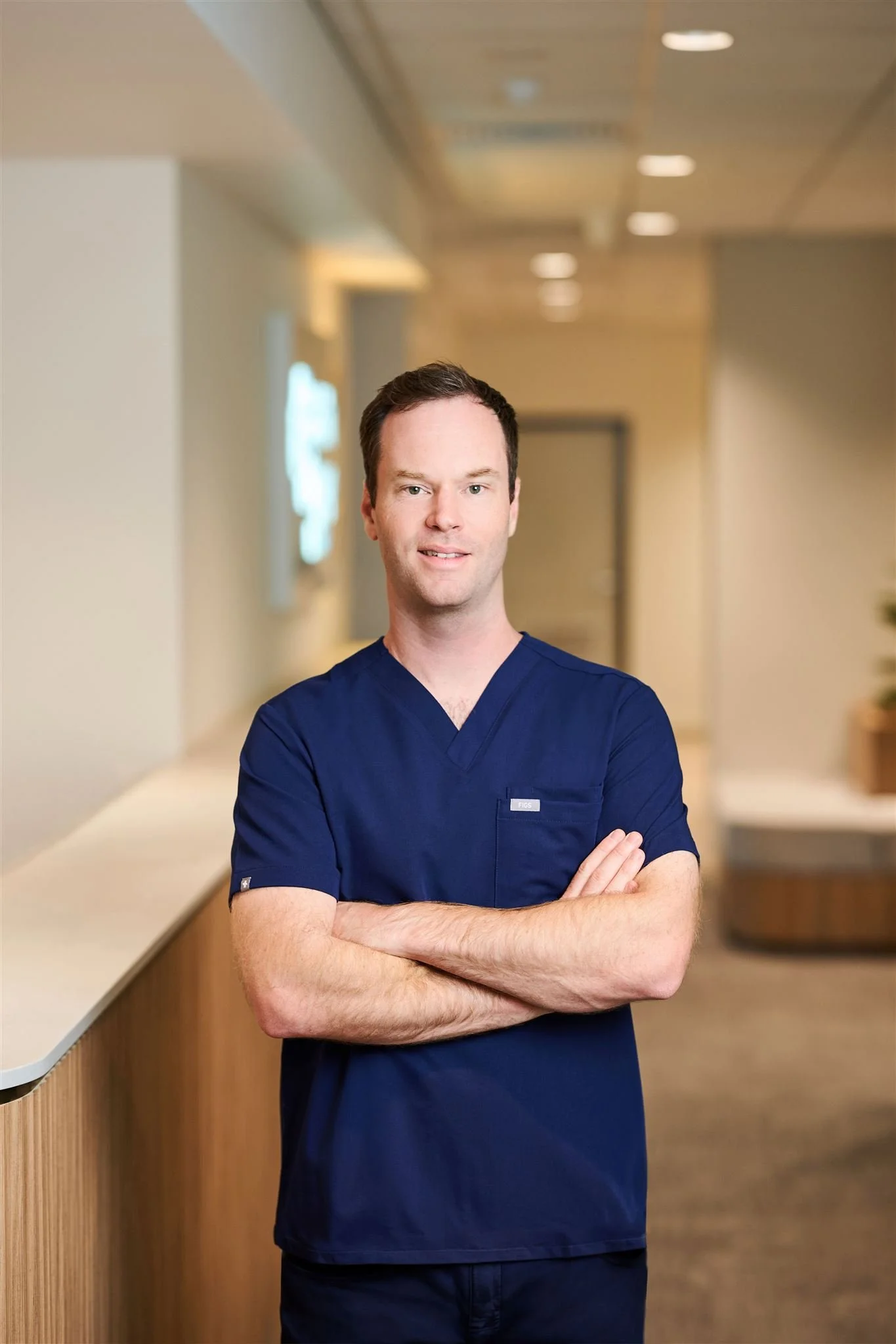 A smiling man in blue medical scrubs standing in a hospital corridor with arms crossed.