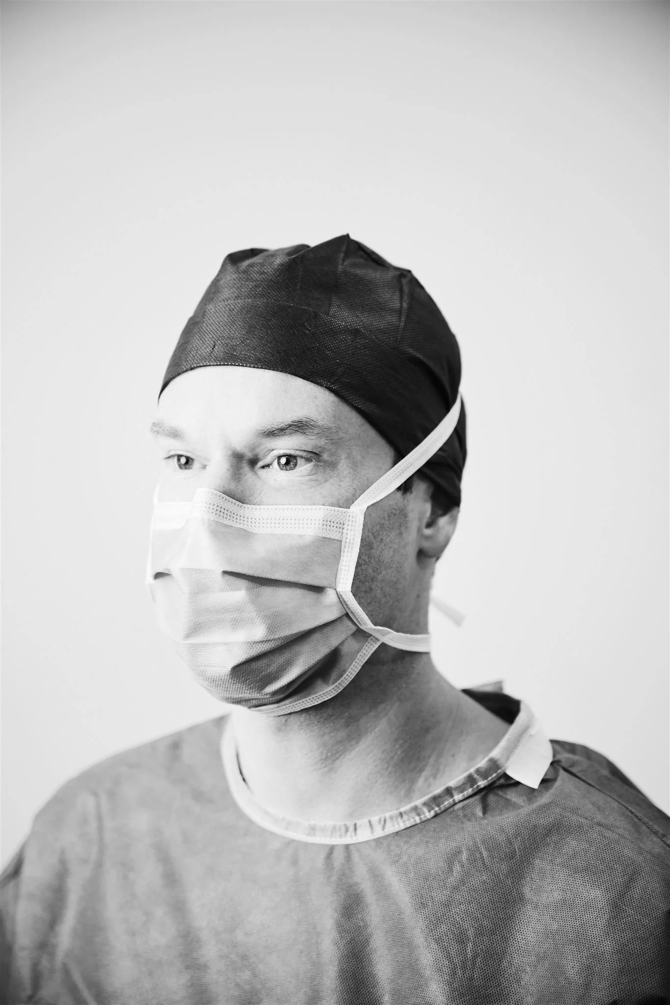 Black and white photo of a male doctor wearing a surgical mask, cap, and scrub top, looking to the right.