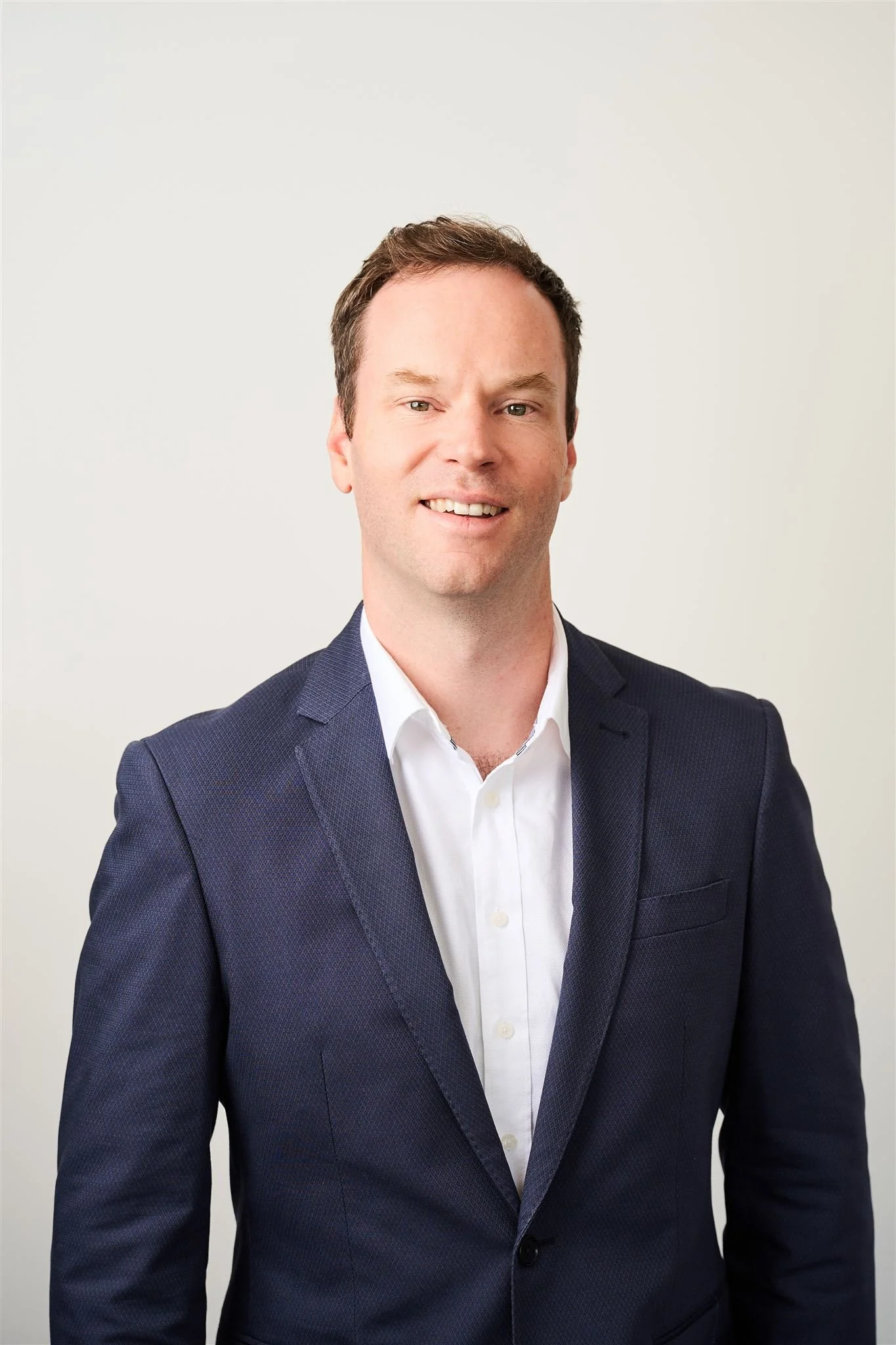 A man in a navy blue suit and white shirt smiling against a plain light background.