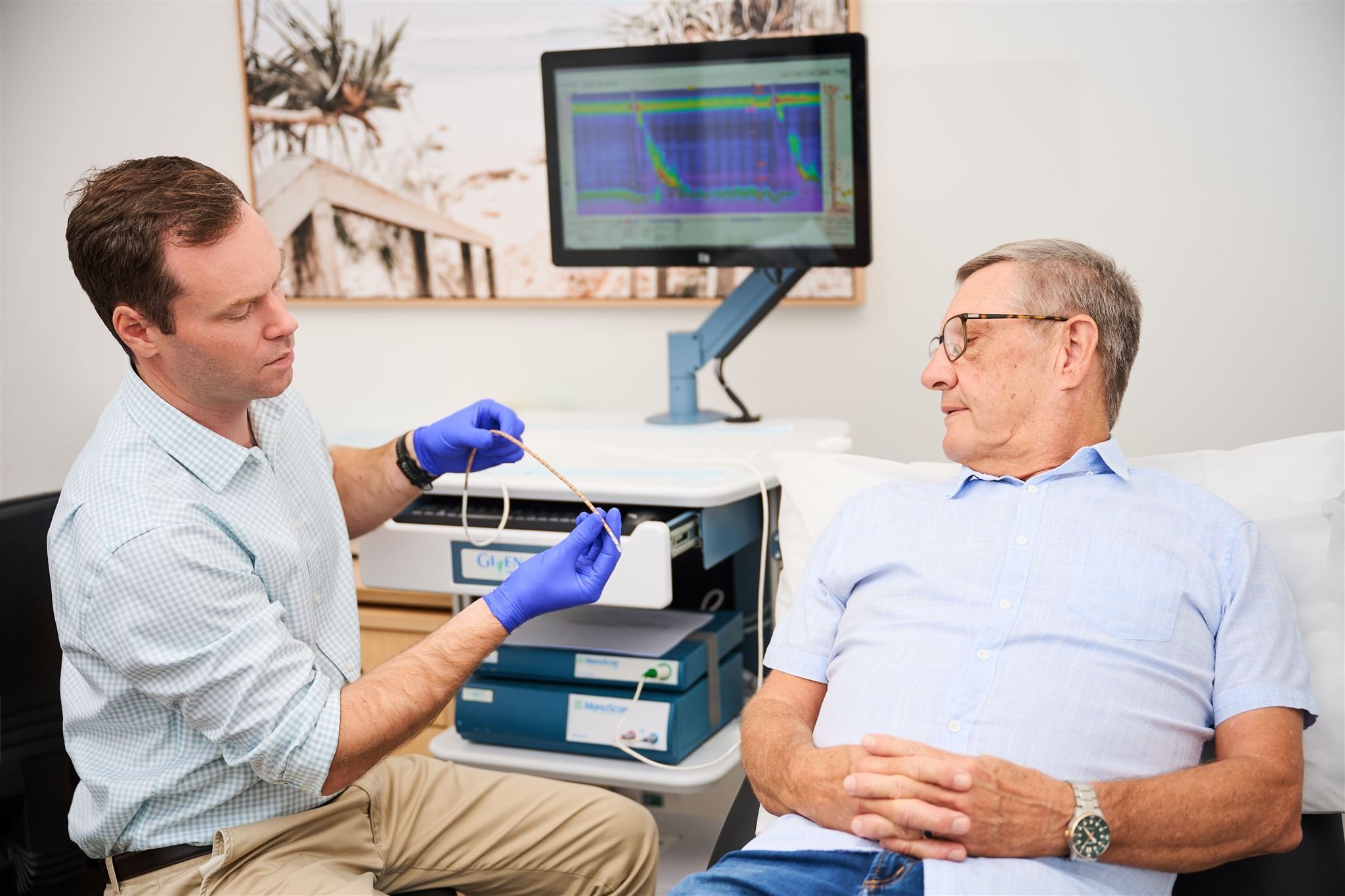 A doctor is testing an elderly man's heart with medical equipment in a hospital room. The man is sitting on a bed with his arms crossed, and a monitor displaying heart data is in the background.