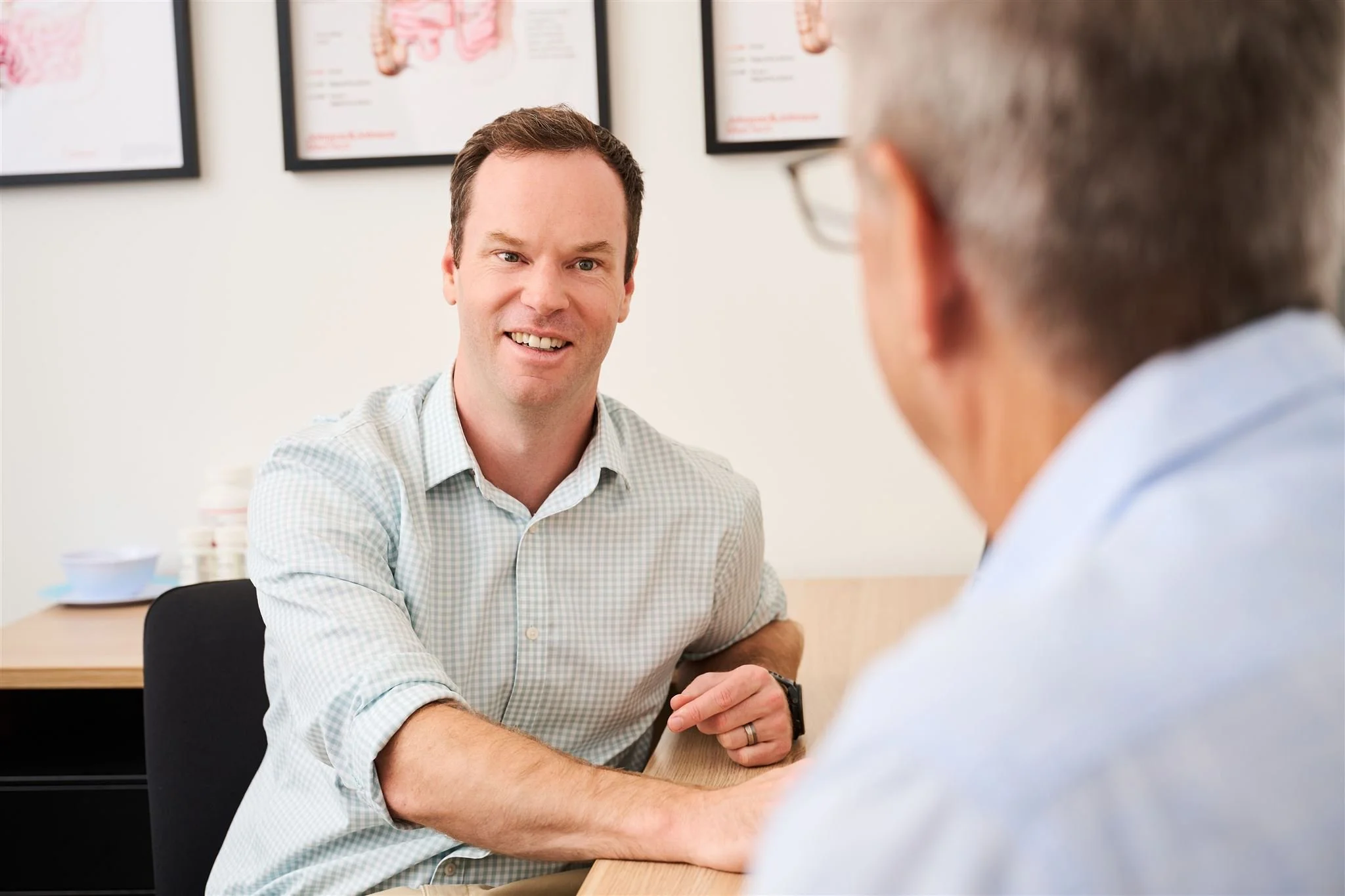 Two men having a conversation in an office, with framed artwork on the wall behind them and a desk with documents and a bowl in the background.