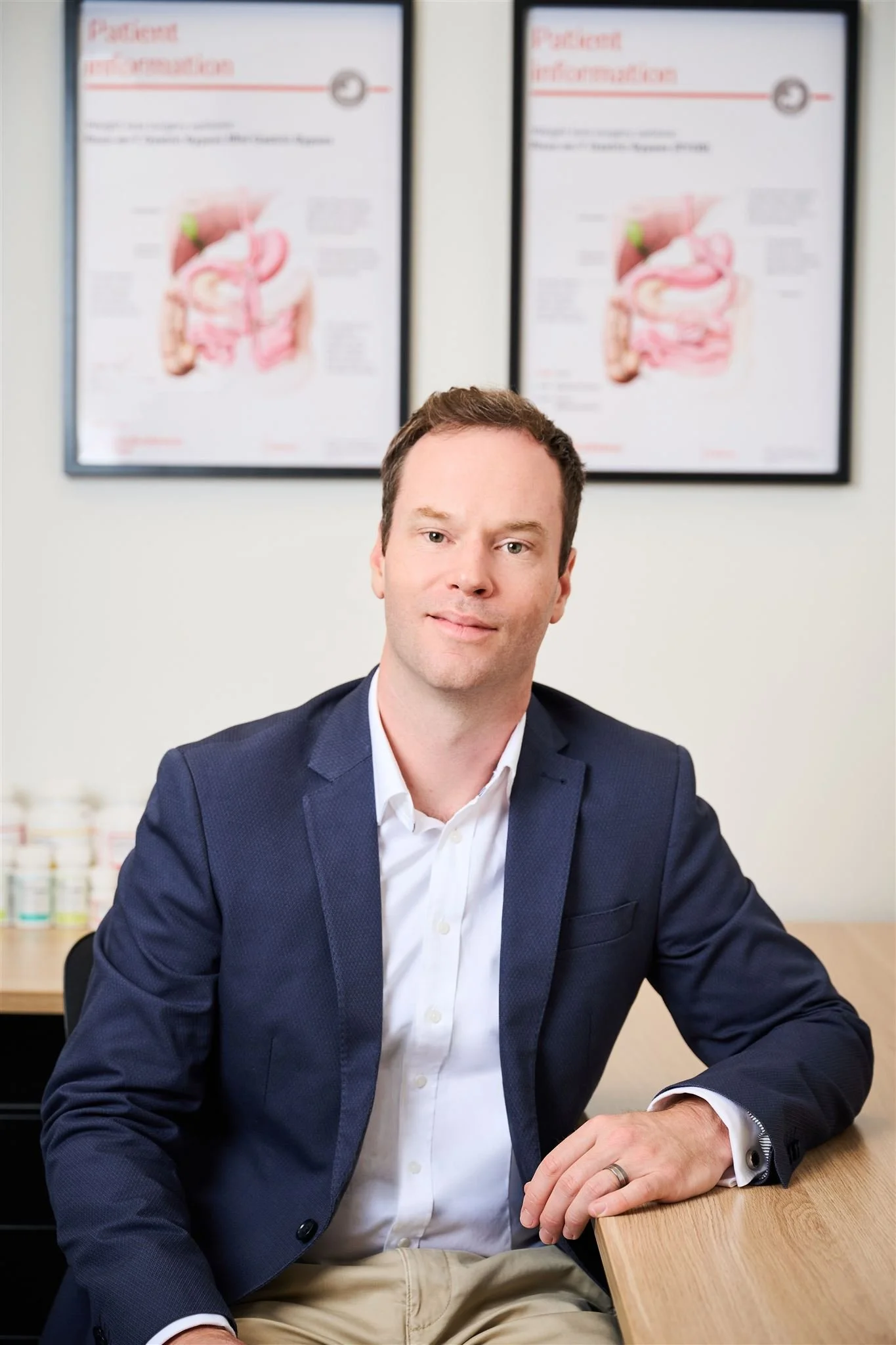 A man dressed in a suit sitting at a desk in an office. Behind him are two medical diagrams labeled 'Patient Information.'