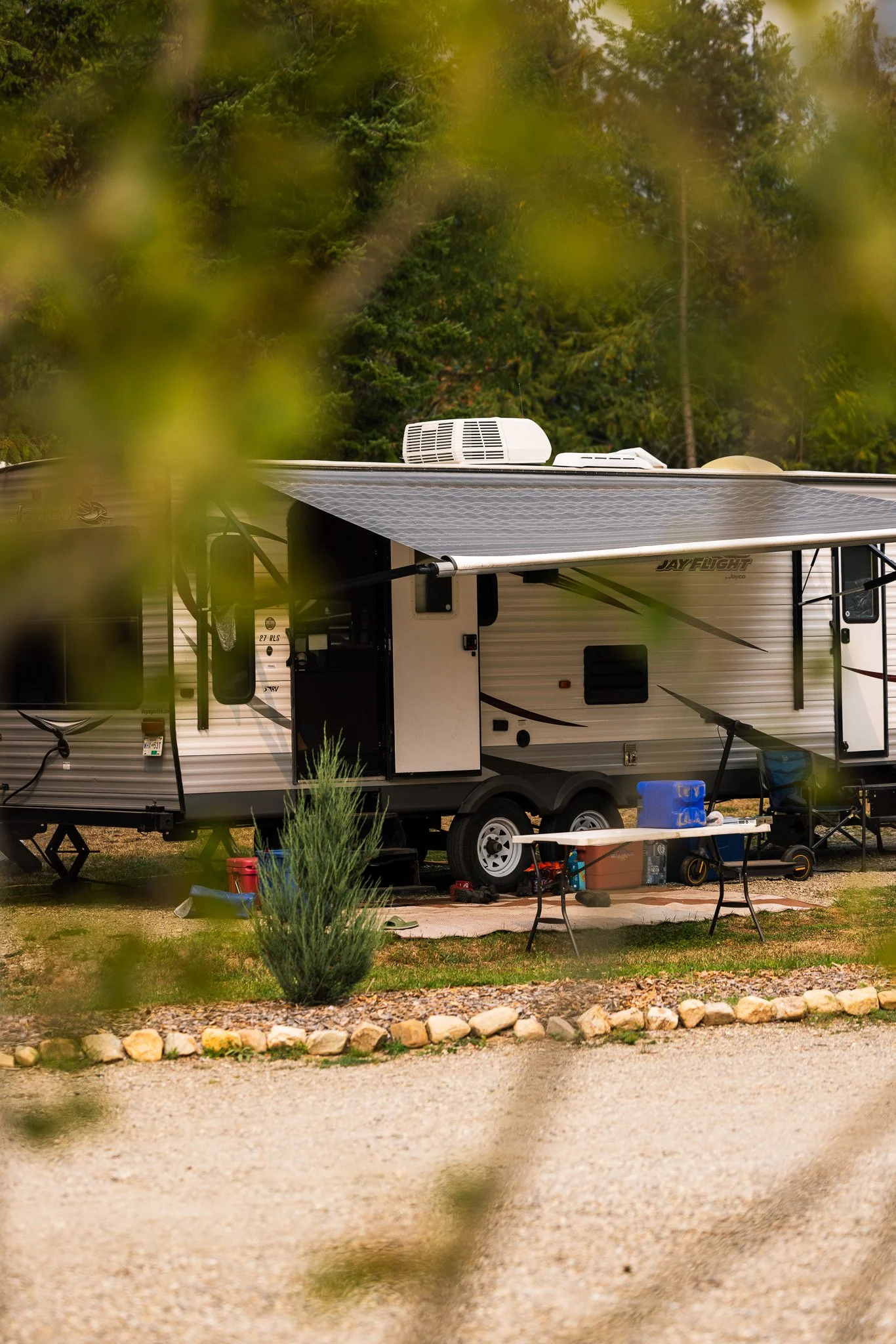 A travel trailer with a solar panel on the roof, parked in a wooded area with trees in the background and a small outdoor setup including a table, chairs, and various items.