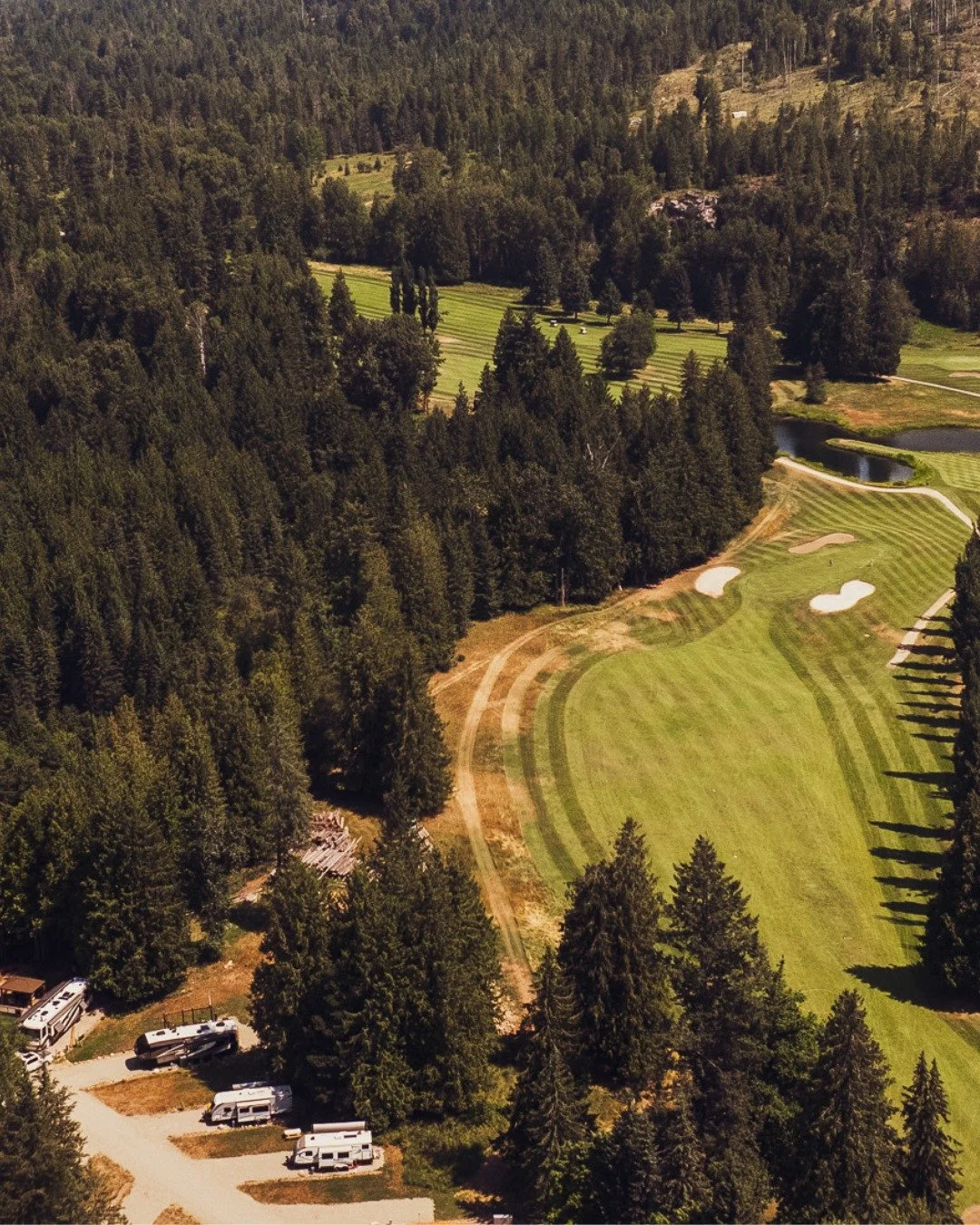 Aerial view of a golf course surrounded by dense forest with several sand traps, water hazards, and a parking area with RVs at the bottom left.